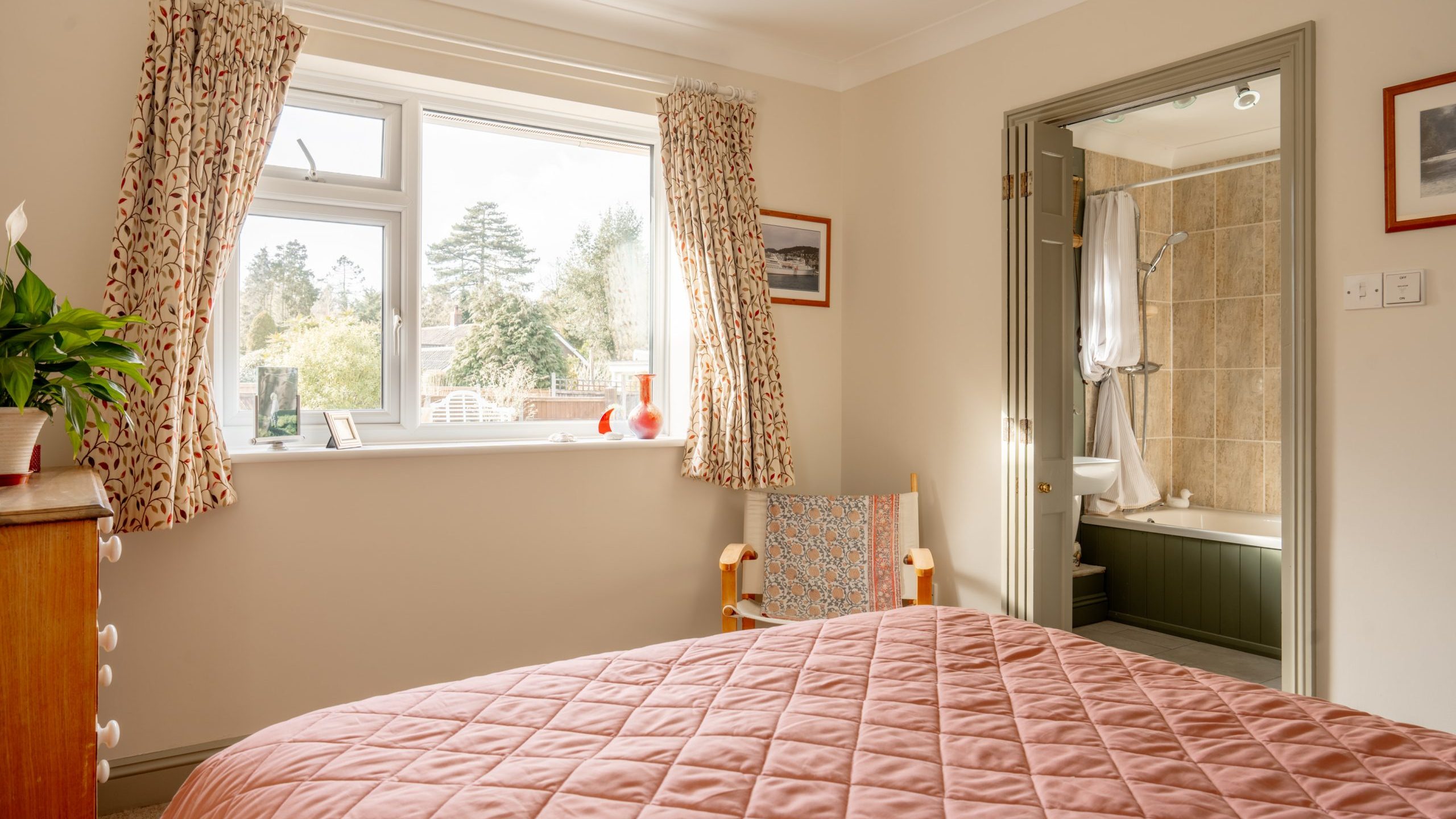 A cozy bedroom with a pink quilted bedspread. There's a wooden dresser with a plant on top, and a window with patterned curtains showing a view of trees. An open door leads to a bathroom with a bathtub and tan tiles. A chair sits by the window.