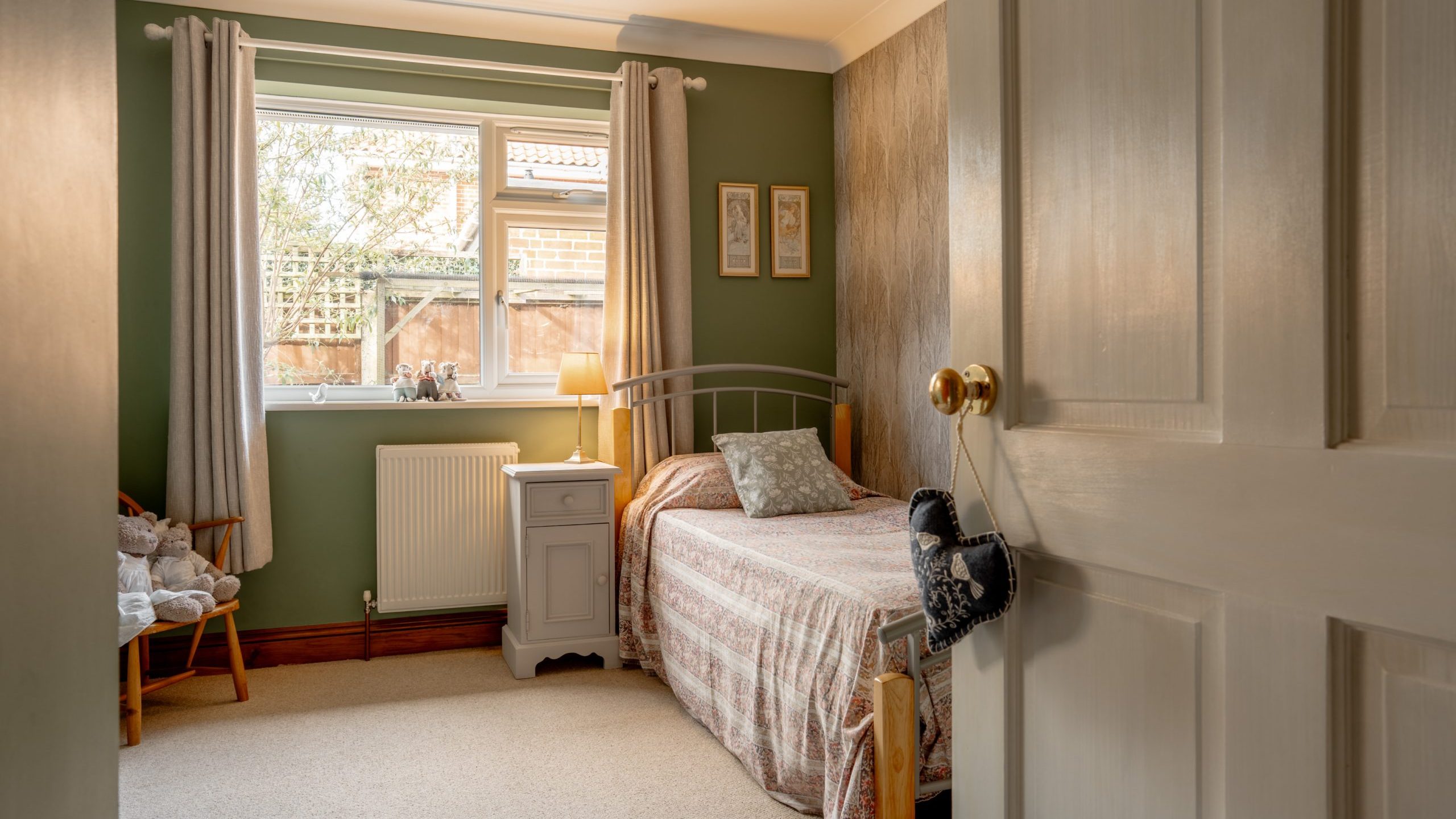 A cozy bedroom with a single bed covered in a floral quilt. The walls are green, and there’s a window letting in natural light. A nightstand with a lamp is beside the bed, and a small chair with stuffed toys sits nearby. The door is slightly open.