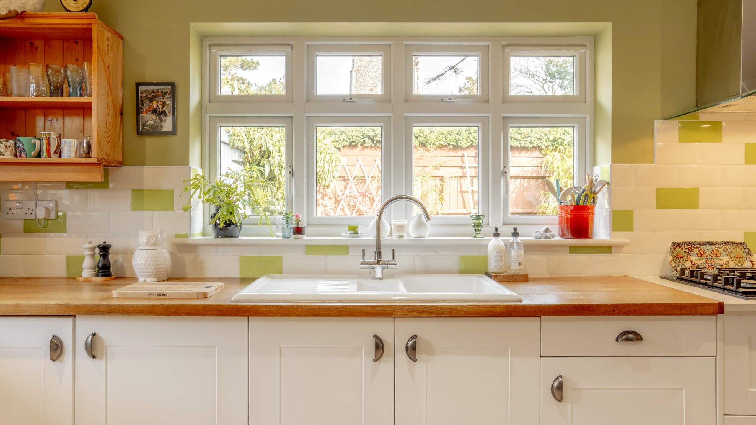 A bright kitchen with green walls and a large window above a white sink. White cabinets and a wooden countertop are present, decorated with herbs and utensils. The window overlooks a garden with plants and a wooden fence.