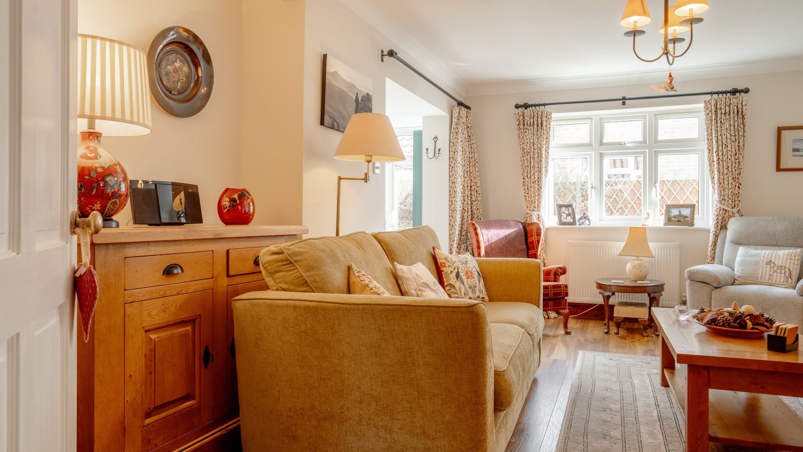 A cozy living room with a sofa, colorful cushions, and wooden furniture. Soft lighting from a lamp and chandelier creates a warm atmosphere. Large window with floral curtains allows natural light. Decorative items are placed on shelves and a coffee table.