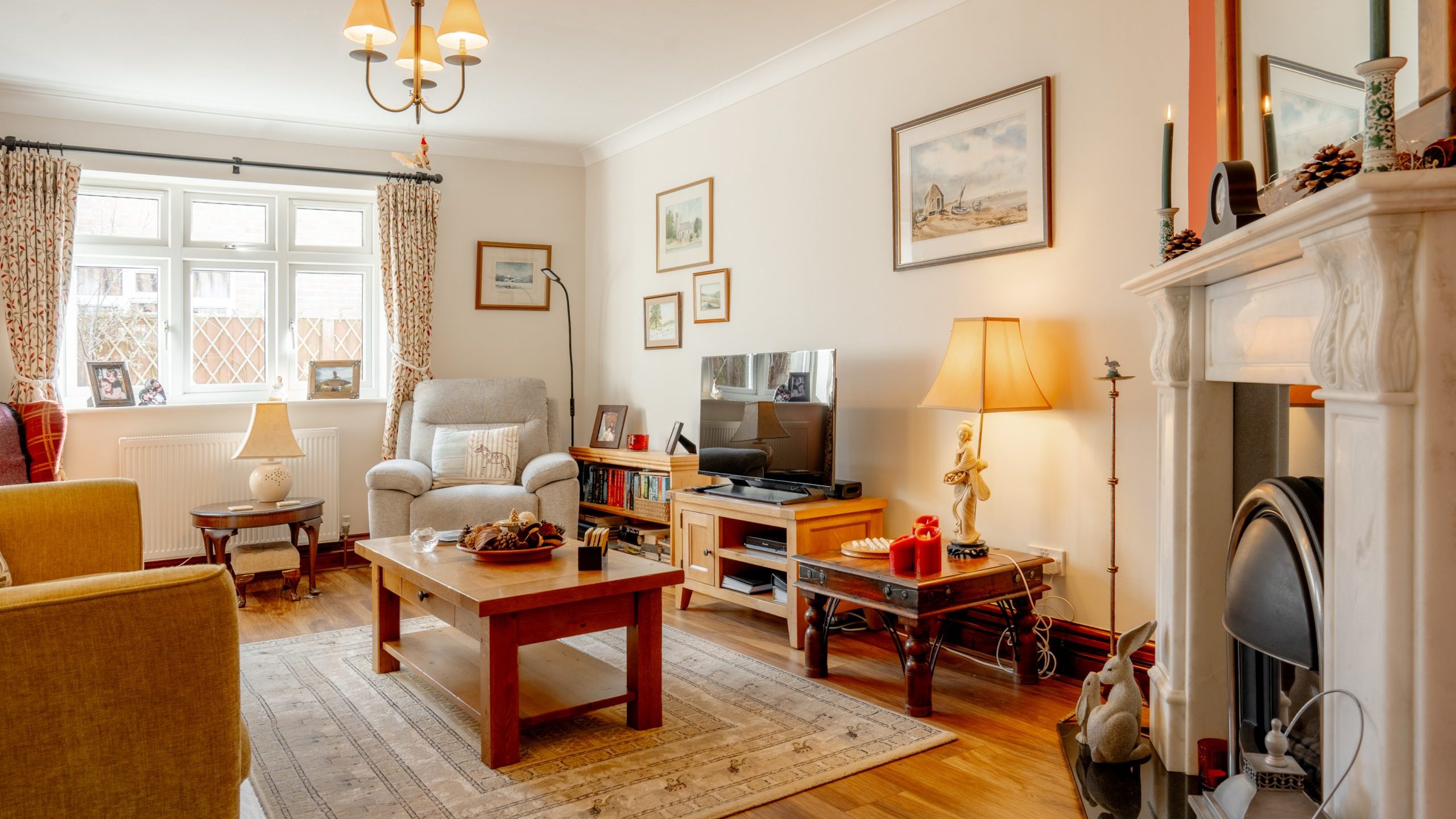 A cozy living room with a beige armchair, yellow sofa, and wooden coffee table on a patterned rug. A TV sits on a wooden stand. The walls are adorned with framed artwork. A fireplace with candles and decorations is on the right, and a large window offers natural light.