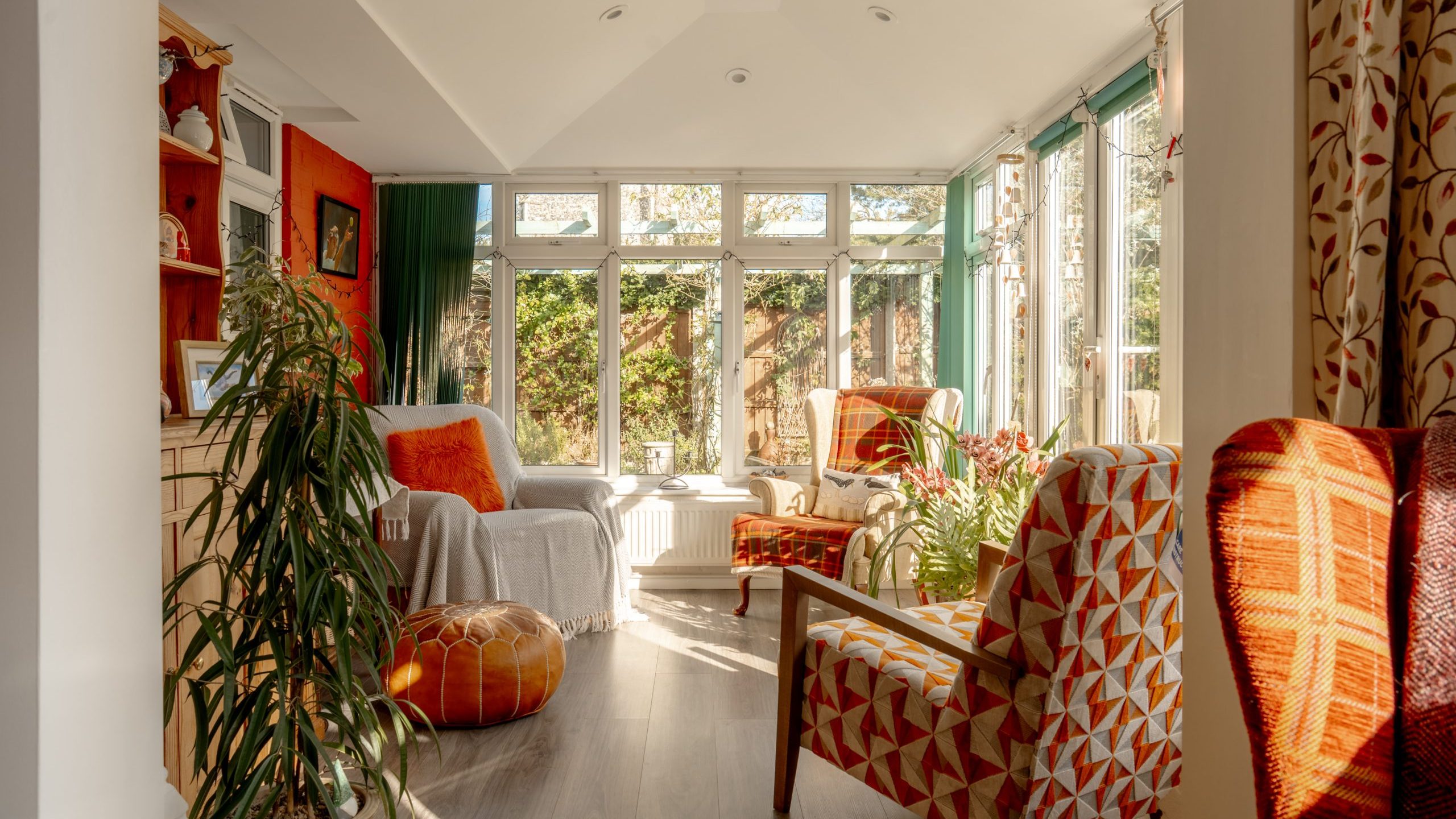 A cozy sunlit room with a large window and garden view. There are patterned orange chairs, a wooden floor, potted plants, and a white side table. The walls are a mix of orange and white, enhancing the warm atmosphere.