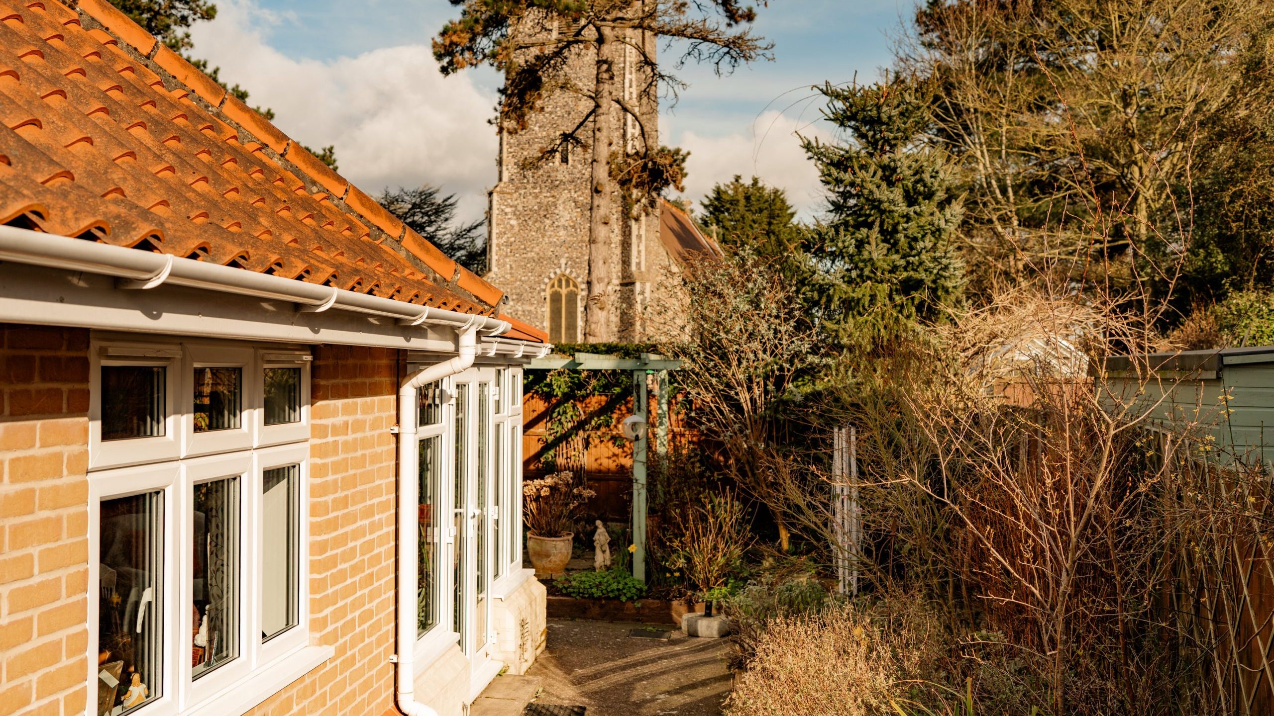 View of a brick house with a red-tiled roof, surrounded by green and dry vegetation, under a partly cloudy sky. In the background, a stone church tower rises, partially obscured by tall trees.