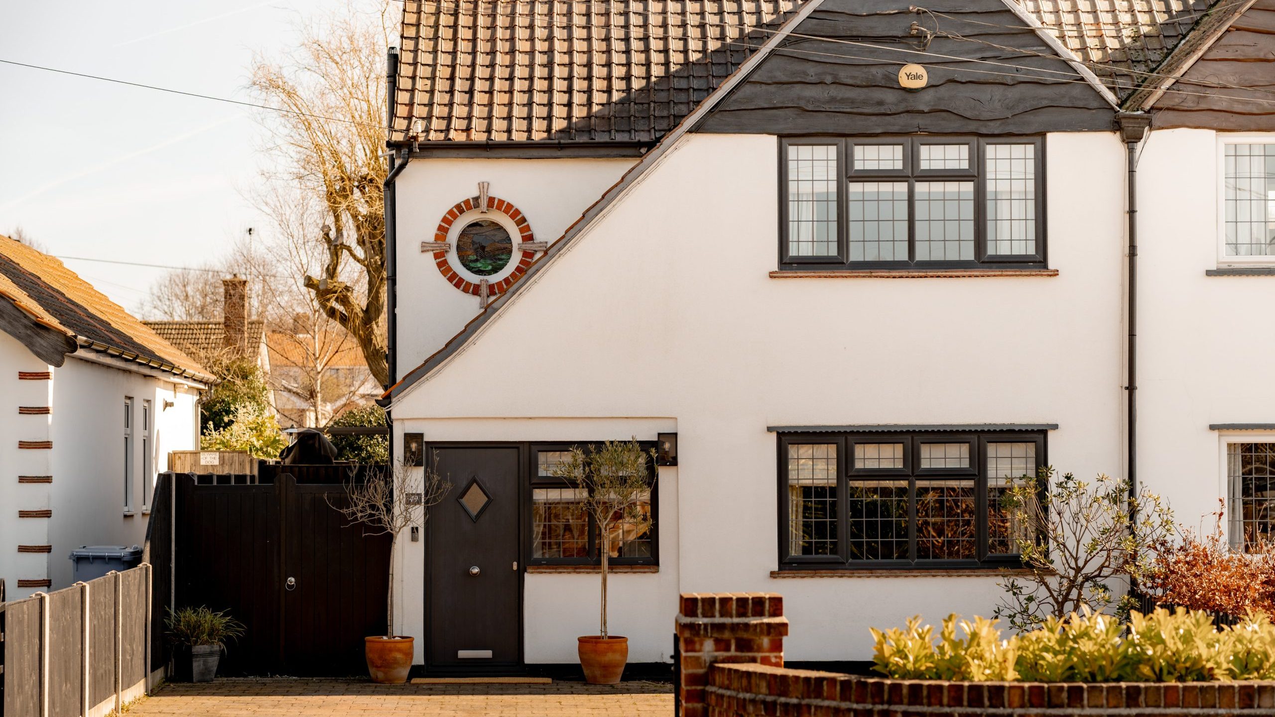 A two-story house with white walls and dark brown trim, featuring a circular window and a central front door. Two potted plants flank the entrance. A brick driveway and a small garden are in front. Another house is partially visible on the left.