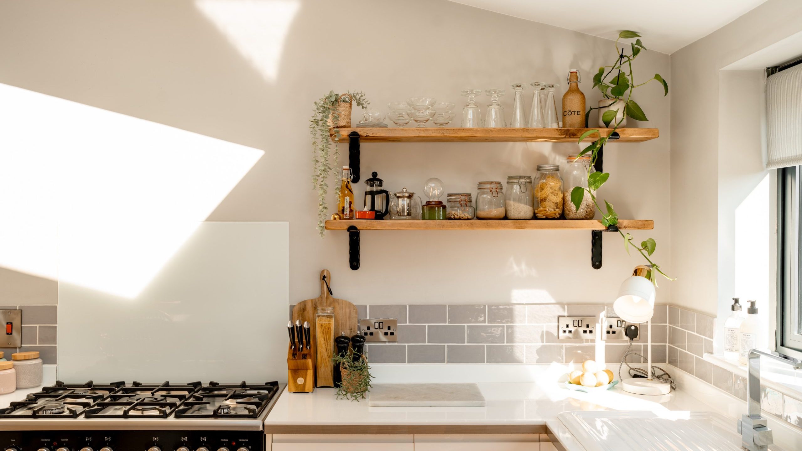 A bright, modern kitchen with light-colored countertops and a gas stove on the left. Wooden shelves above hold glassware, plants, and jars. Subtle gray tiles adorn the backsplash. A window on the right brings in natural light.