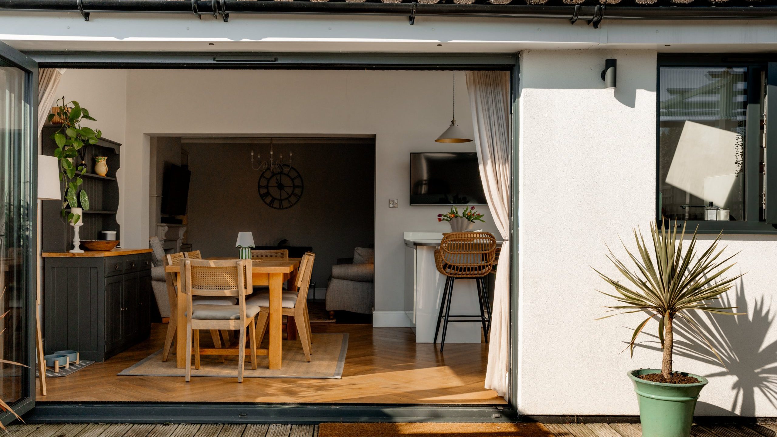 Open sliding door reveals an indoor dining and living area with wooden furniture and a large clock on the wall. The space is bright, featuring a dining table, bar stools at a counter, and a potted plant outside on a wooden deck.