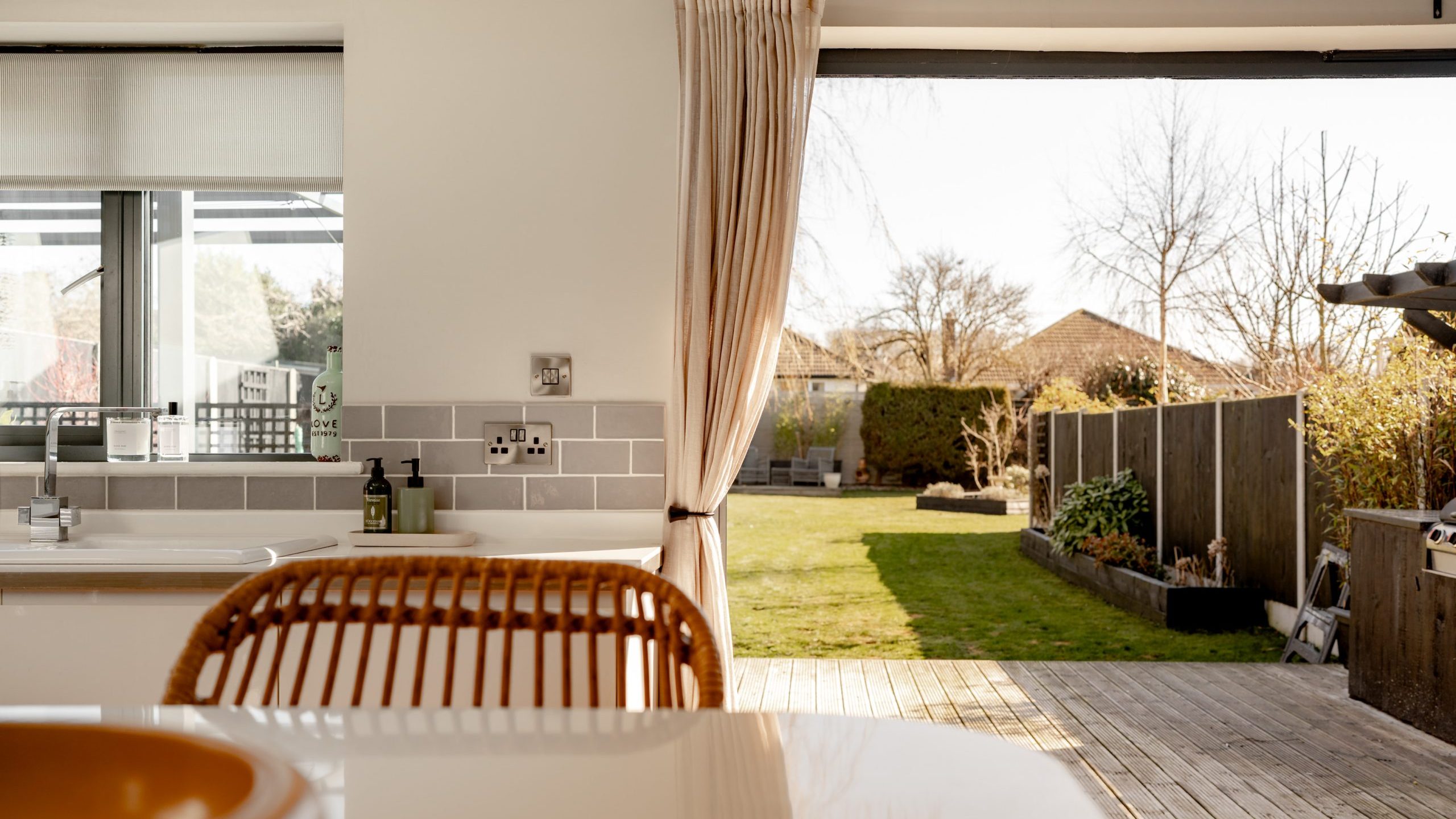 Open kitchen view with a wooden countertop and wicker chair, facing sliding doors leading to a sunlit garden. The garden has a wooden deck, green lawn, and fenced sides. Bright, natural light filters through the space.