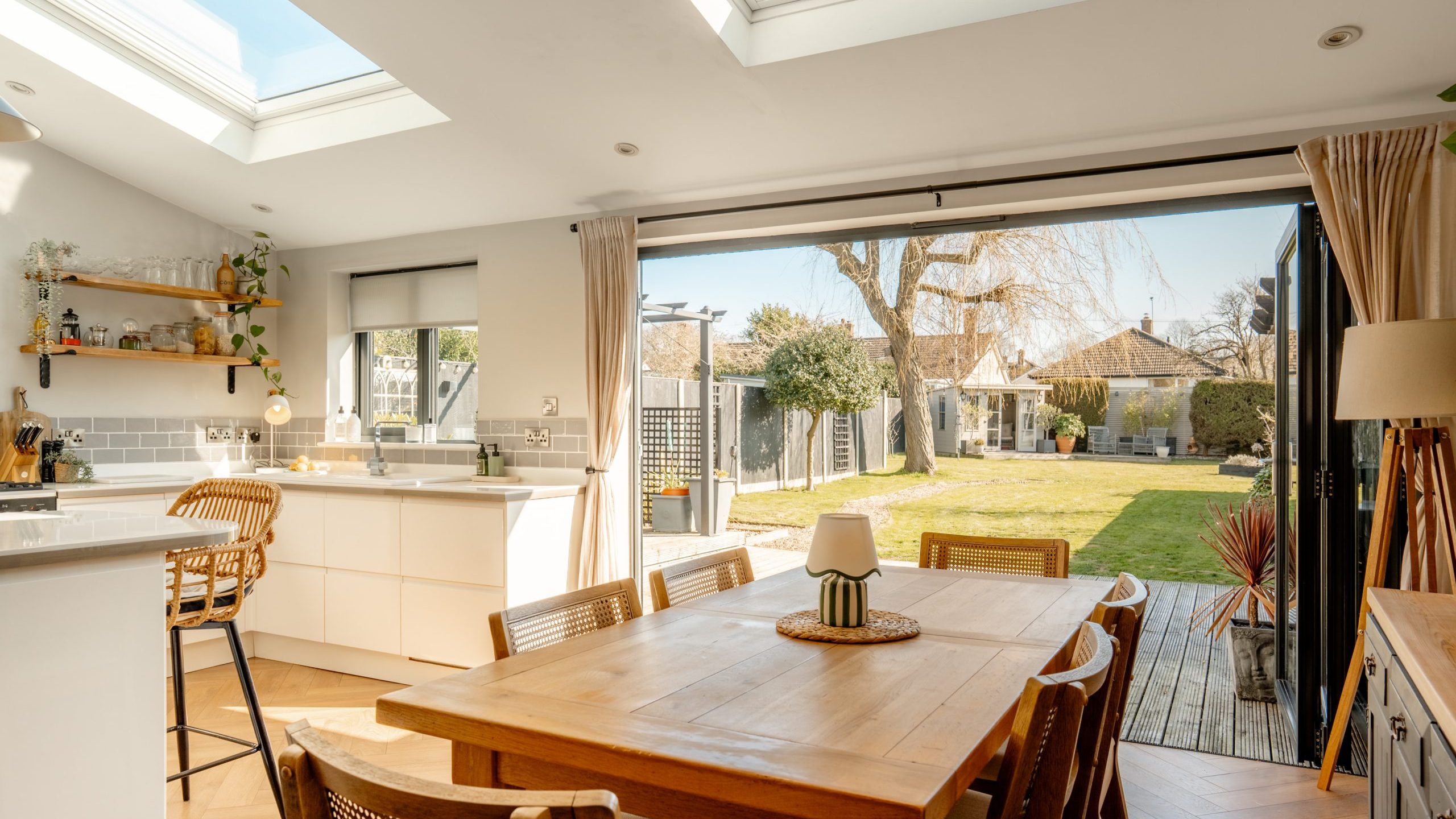 A bright, modern kitchen and dining area with skylights, wooden table, and wicker chairs. Large patio doors open to a garden with a lawn, trees, and houses in the background. White cabinetry and light wood floors create an airy atmosphere.