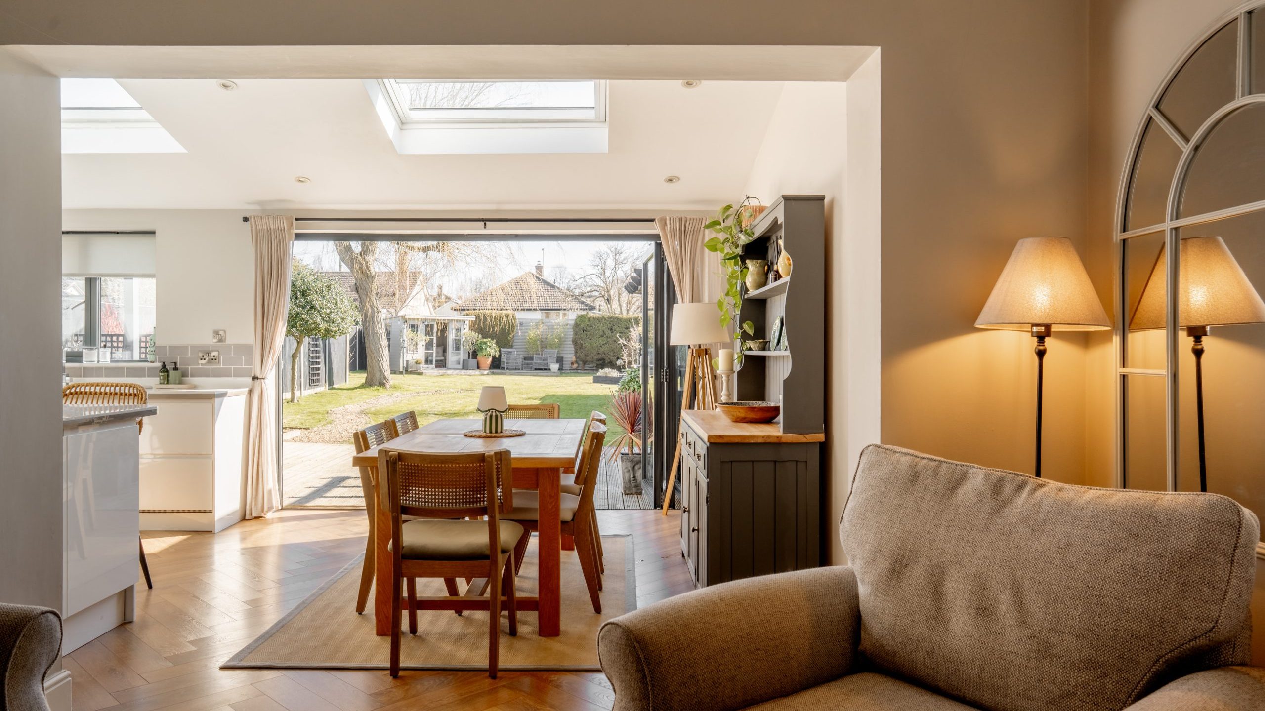 Bright, open-plan living space with a cozy armchair in the foreground, a wooden dining table, and chairs near French doors leading to a sunny garden. Skylights and soft lighting enhance the welcoming atmosphere.