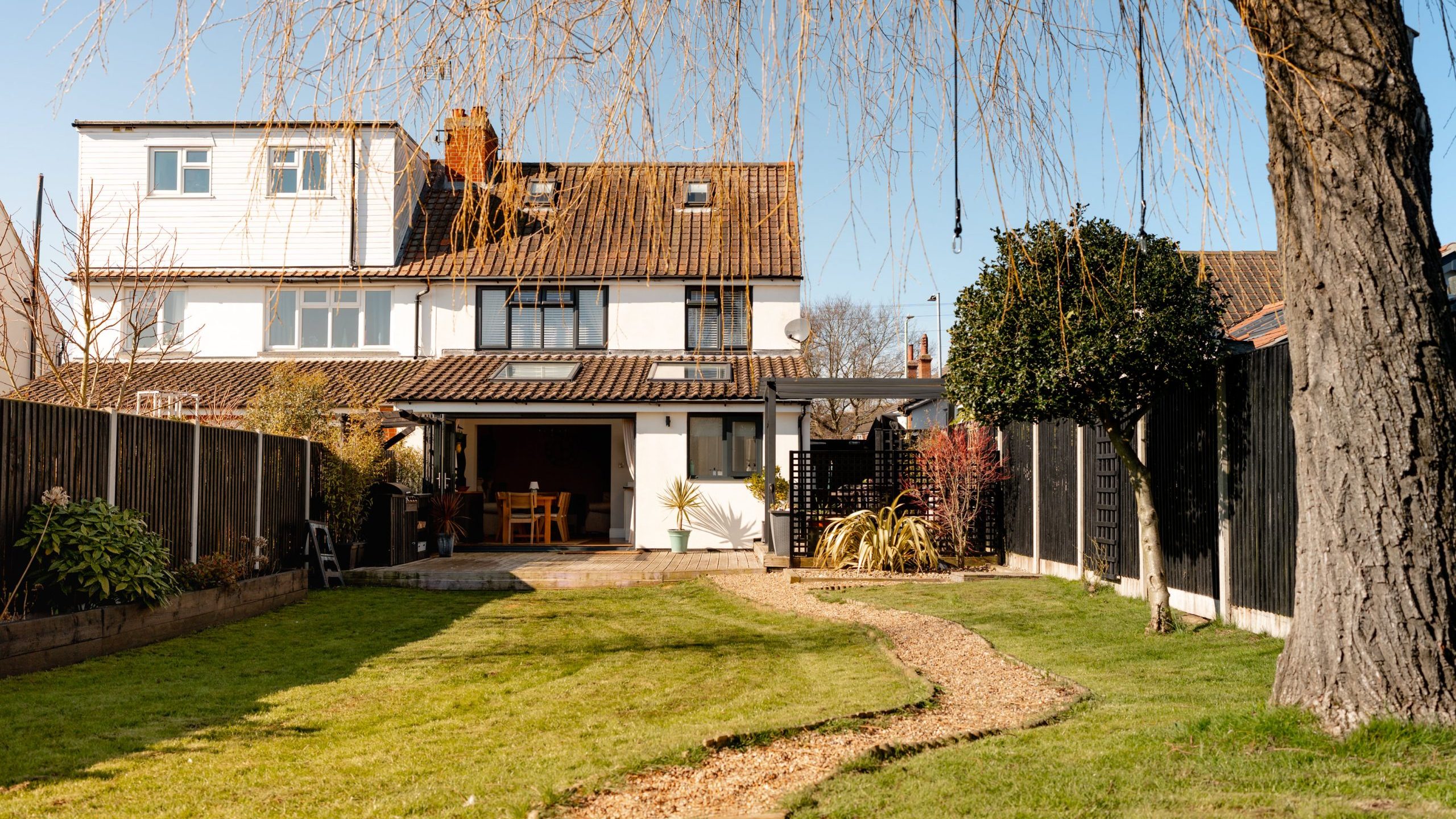A two-story house with a sloped roof, large windows, and an extended patio. The backyard features a winding gravel path, bordered by green grass and fenced on both sides. A tall tree stands on the right, with a clear blue sky overhead.
