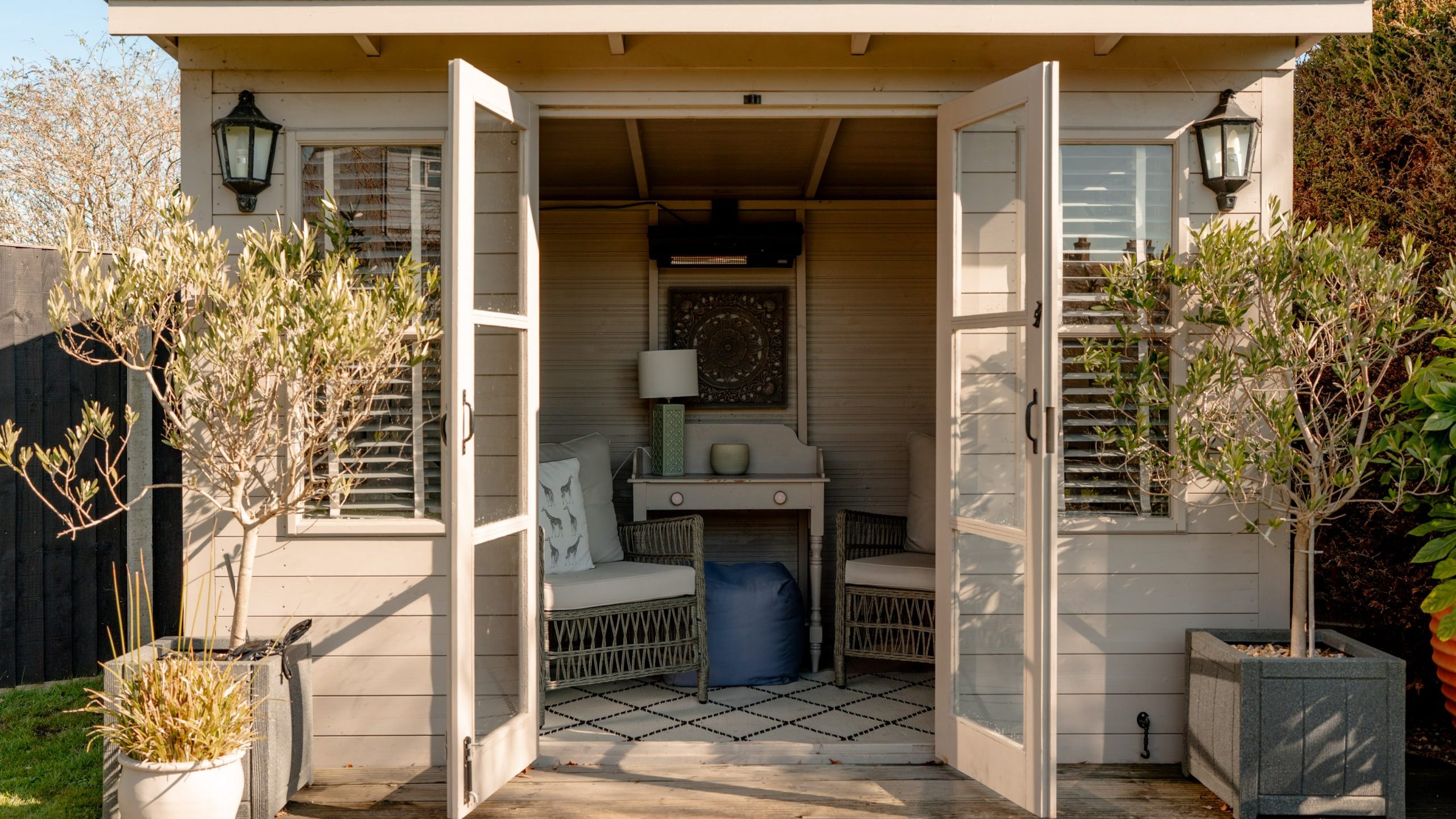A cozy garden shed with open double doors, revealing a small sitting area with two chairs and a table inside. The exterior features lantern lights and potted plants at the entrance. Sunlight illuminates the interior, highlighting a decorative wall piece.