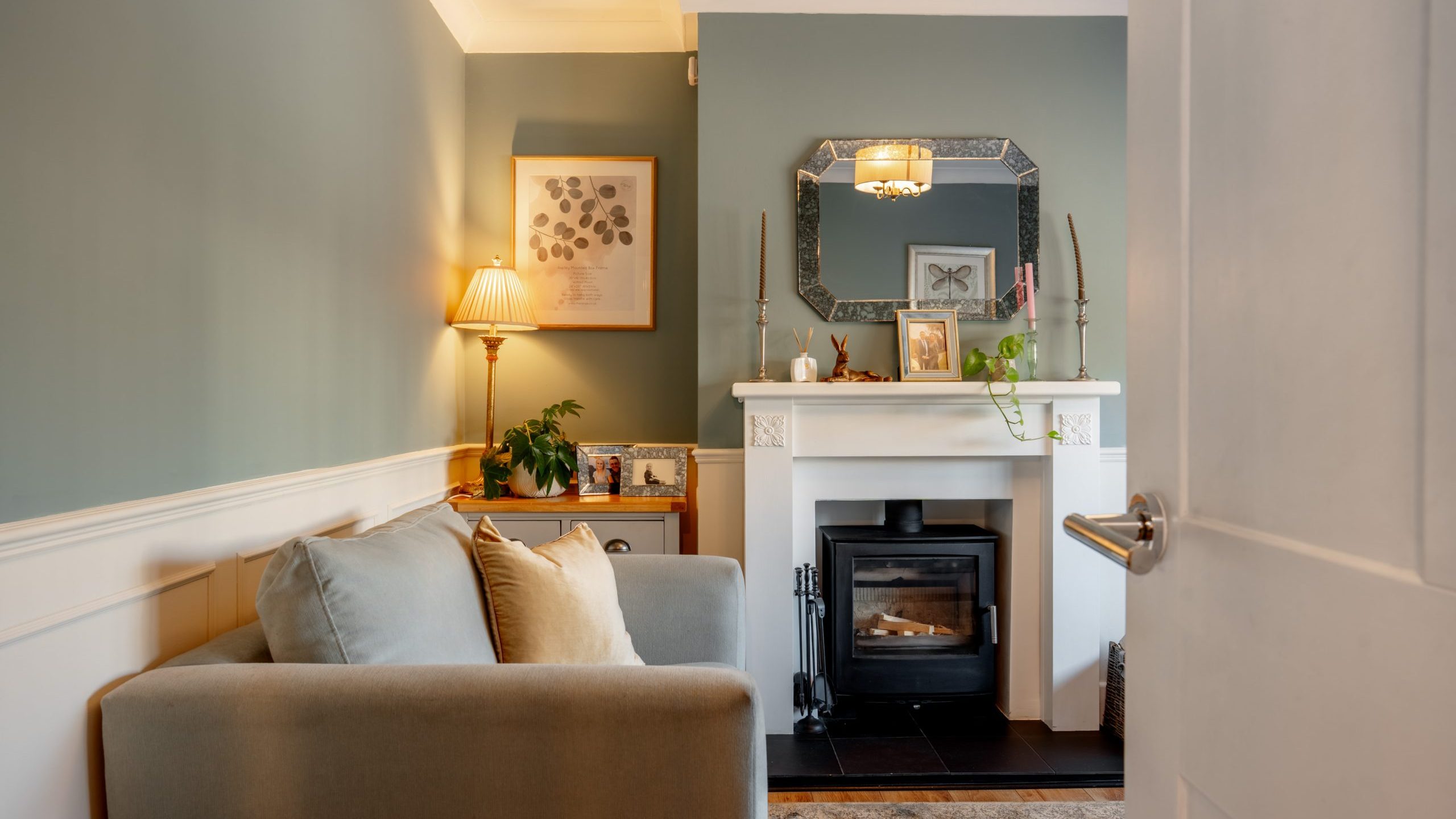 A cozy living room with a gray couch, soft cushions, and a modern fireplace. The walls are painted green and adorned with framed art. A lamp with a beige shade stands beside a plant. A large mirror and candles decorate the mantel.