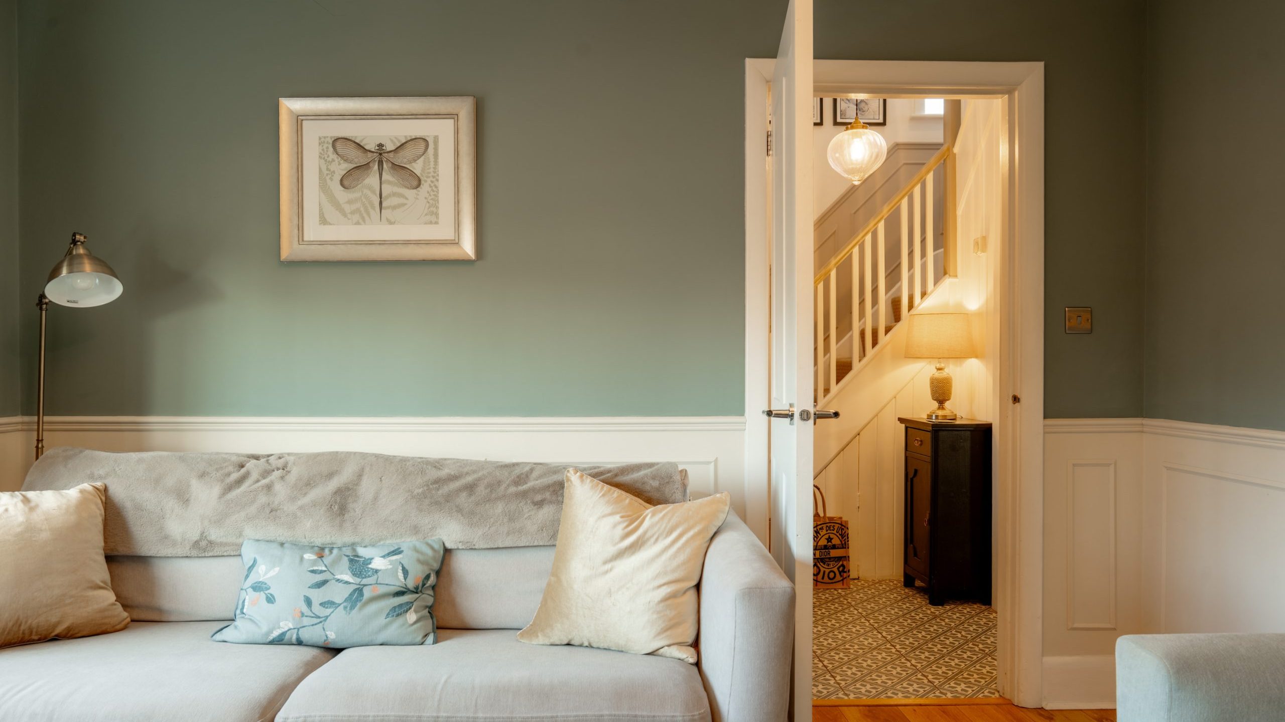 A cozy living room with a light beige sofa adorned with blue and gold cushions. A standing lamp is beside it. The wall is painted green with a butterfly artwork. An open door reveals a lit hallway with a staircase and a lamp on a table.