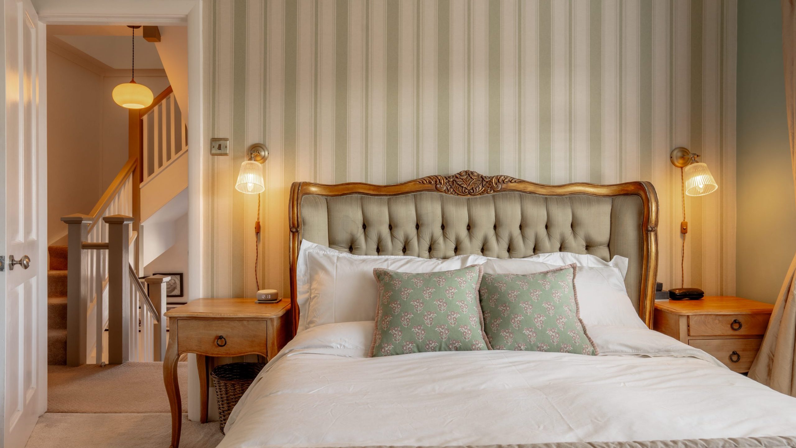 A cozy bedroom with a large bed featuring a tufted headboard and patterned pillows. Wooden side tables with wall-mounted lamps flank the bed. A door opens to a hallway with stairs, softly lit by a hanging lamp. Striped wallpaper decorates the room.