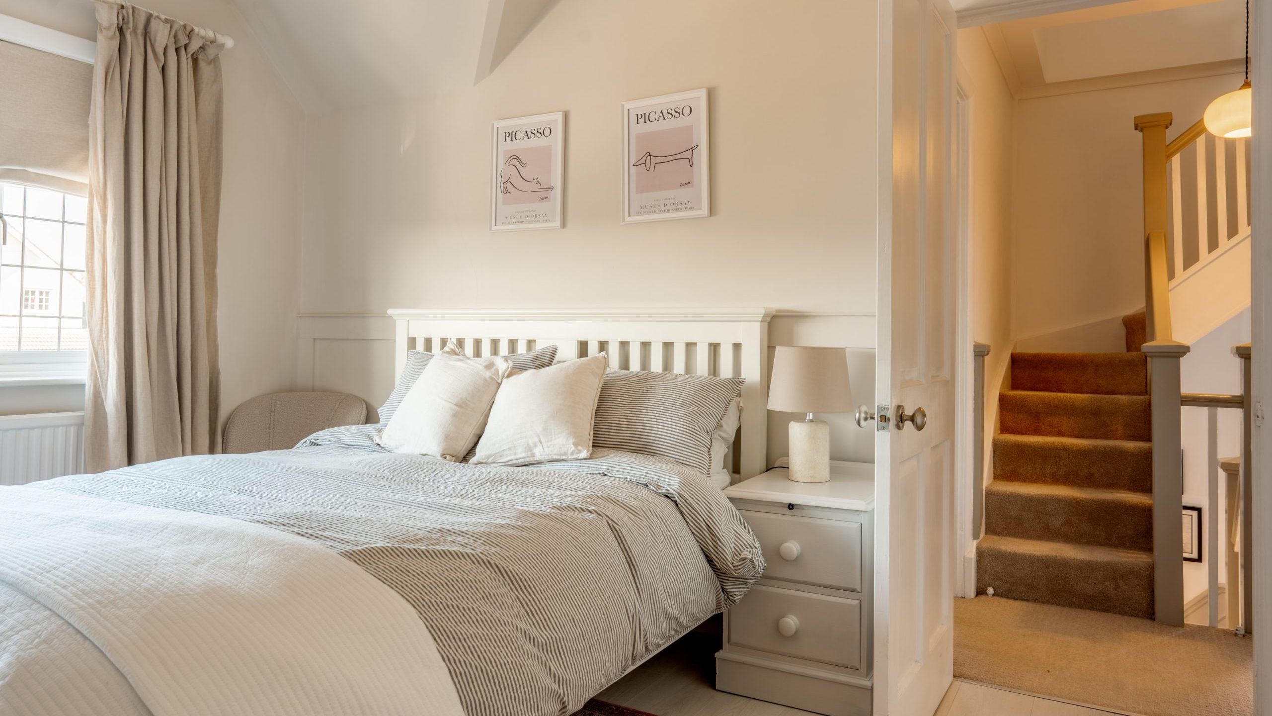A cozy bedroom with a neatly made bed featuring white and gray bedding. Two framed Picasso prints hang above the bed. A bedside table with a lamp sits beside the bed. An open door reveals a staircase leading to another floor.