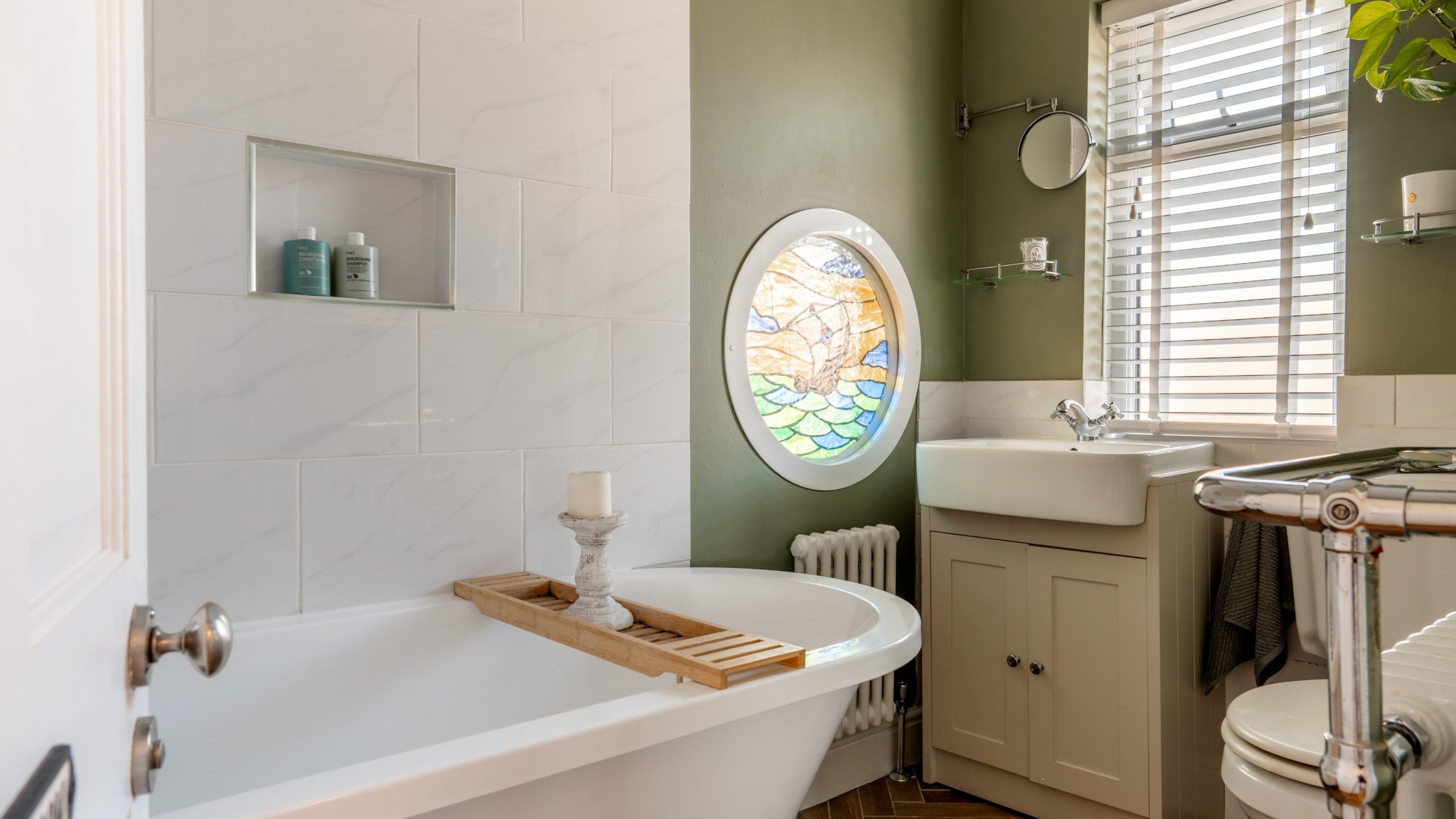 A bathroom with a green accent wall features a white freestanding bathtub with a wooden tray holding a candle. There's a round stained glass window, a sink with a mirror above, and shelves. The floor has a wood pattern.