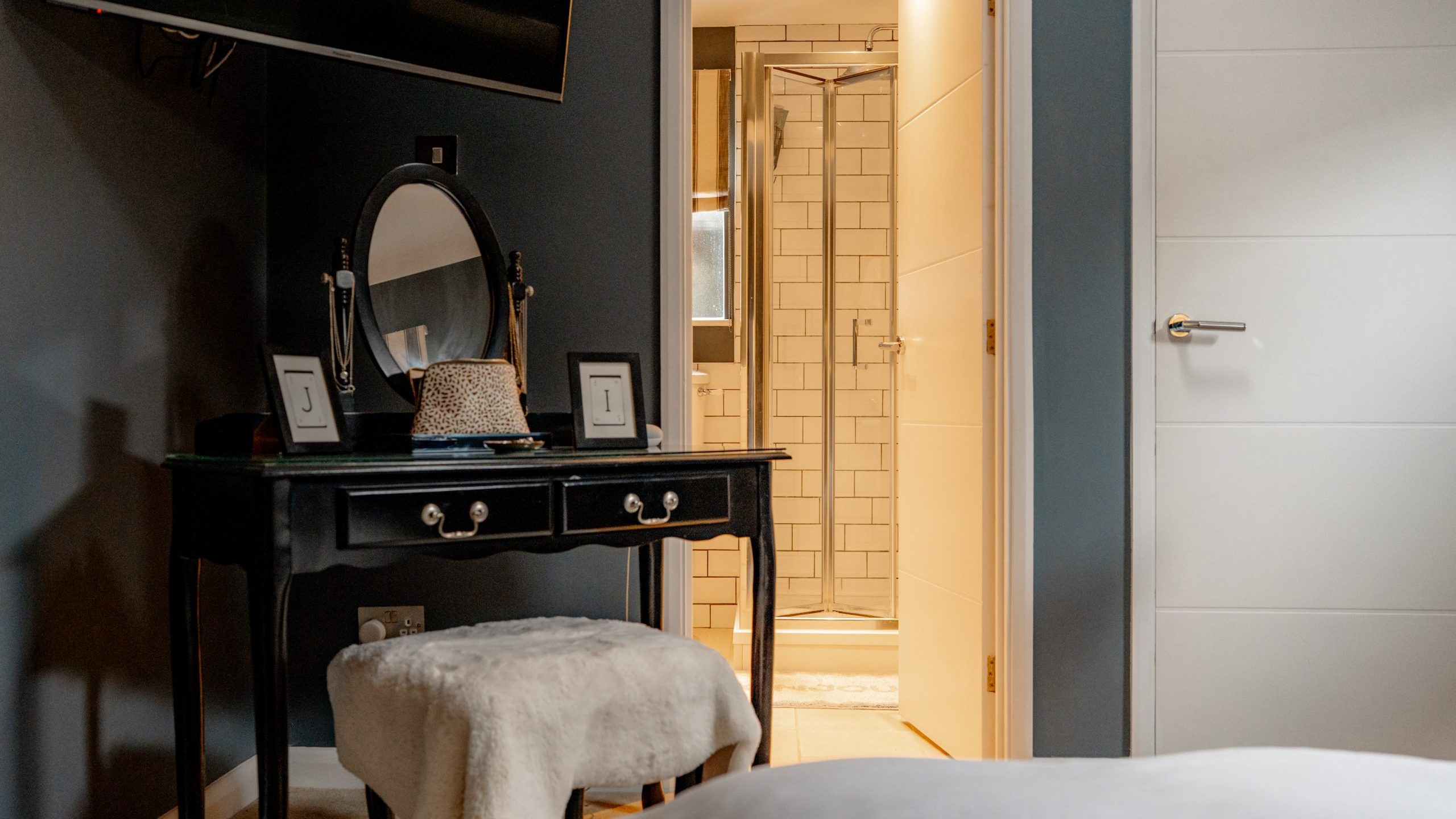 A bedroom features a black vanity with a round mirror, two framed photos, and a hat. A soft stool is placed in front. An open door reveals a bathroom with a shower and white tile. The room is softly lit and has dark walls.