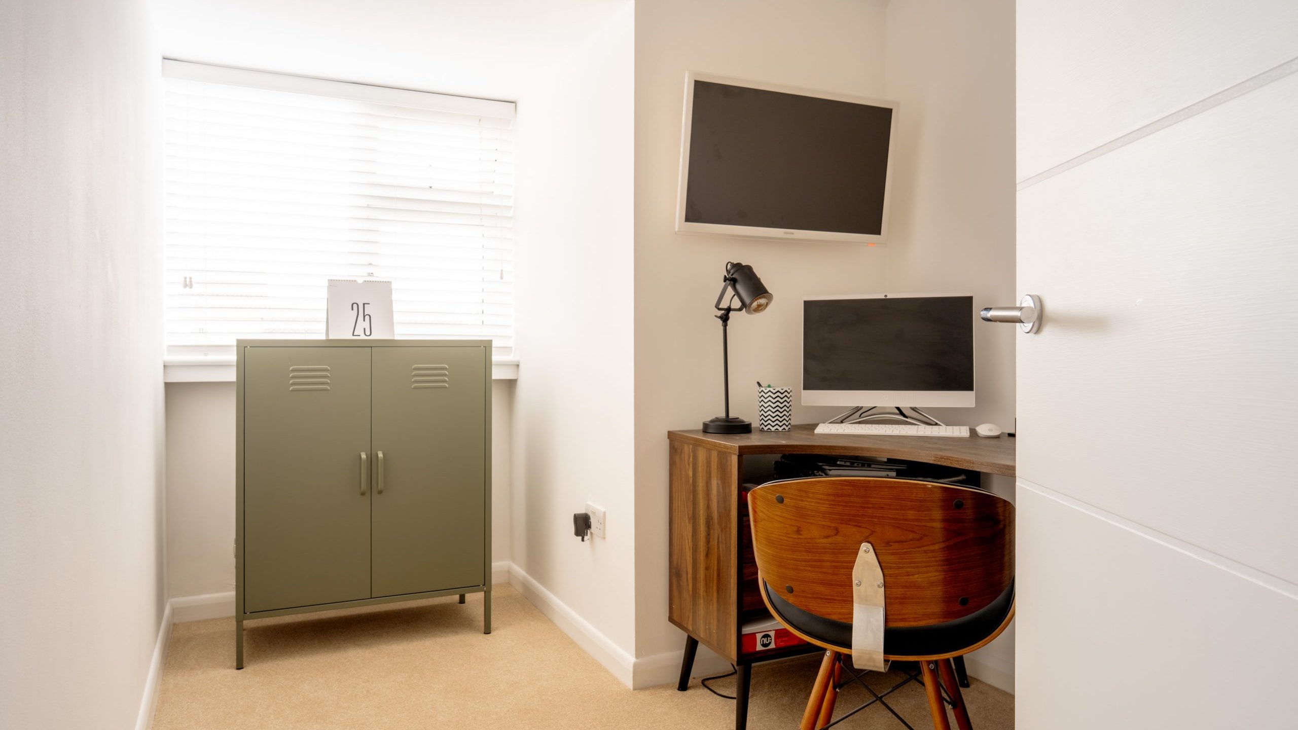 Small home office with a wooden desk and chair, a computer monitor, and a desk lamp. A wall-mounted TV hangs above the desk. A green metal cabinet is against the wall near a window with blinds. White walls and carpeted floor.
