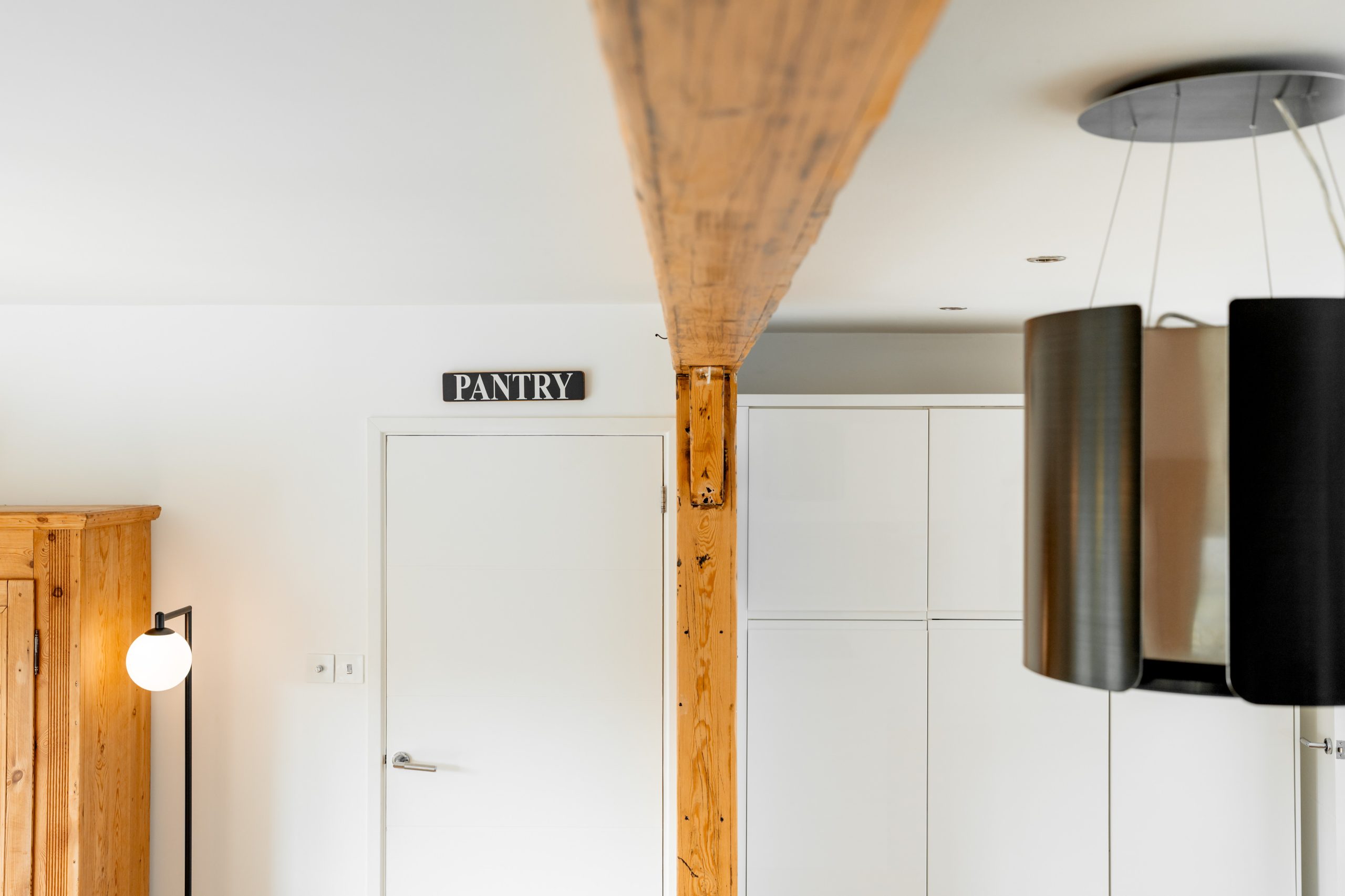 Modern kitchen interior featuring a white pantry door with a black sign, wooden beams, a tall wooden cabinet with a round lamp, and black hanging light fixtures. The space is minimally styled for a clean look.