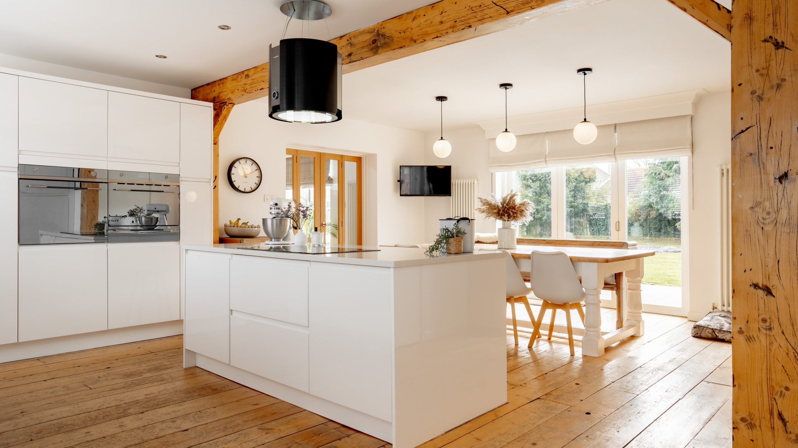 Spacious modern kitchen with wooden beams and light wood flooring. Features white cabinets, a central island with a mixer, wall-mounted clock, dining table with white chairs, and large windows letting in natural light.