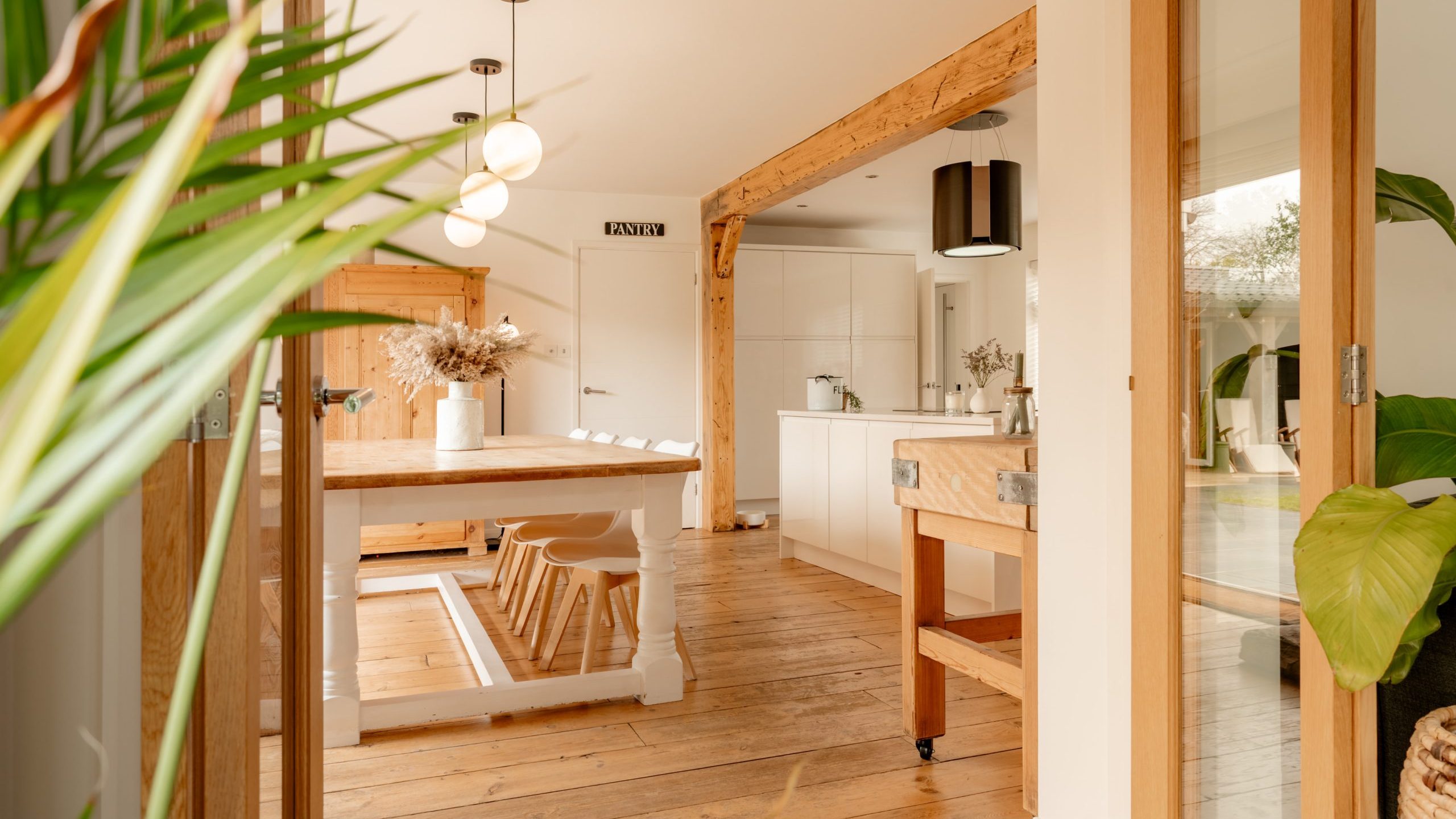 Bright kitchen with wooden floors and beams, featuring a long dining table with stools, hanging pendant lights, and white cabinets. Large houseplants frame the space, adding a touch of greenery. Light streams in through glass doors and windows.