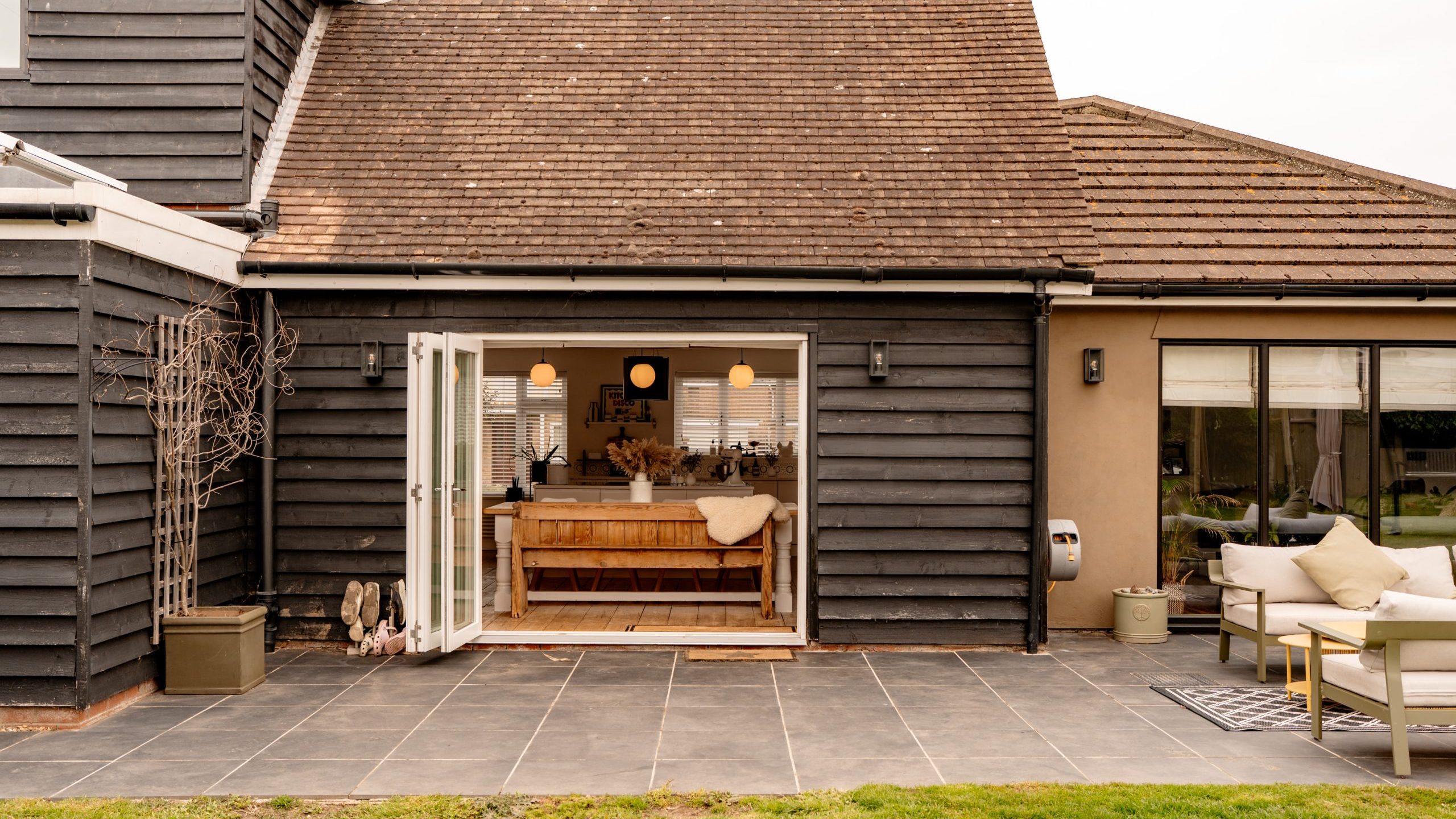 Exterior view of a house with open glass doors leading to a cozy dining area. The house has dark wooden siding and a sloped roof. A patio with gray tiles and grass surrounds the area, and there's outdoor seating with cushions on the right.