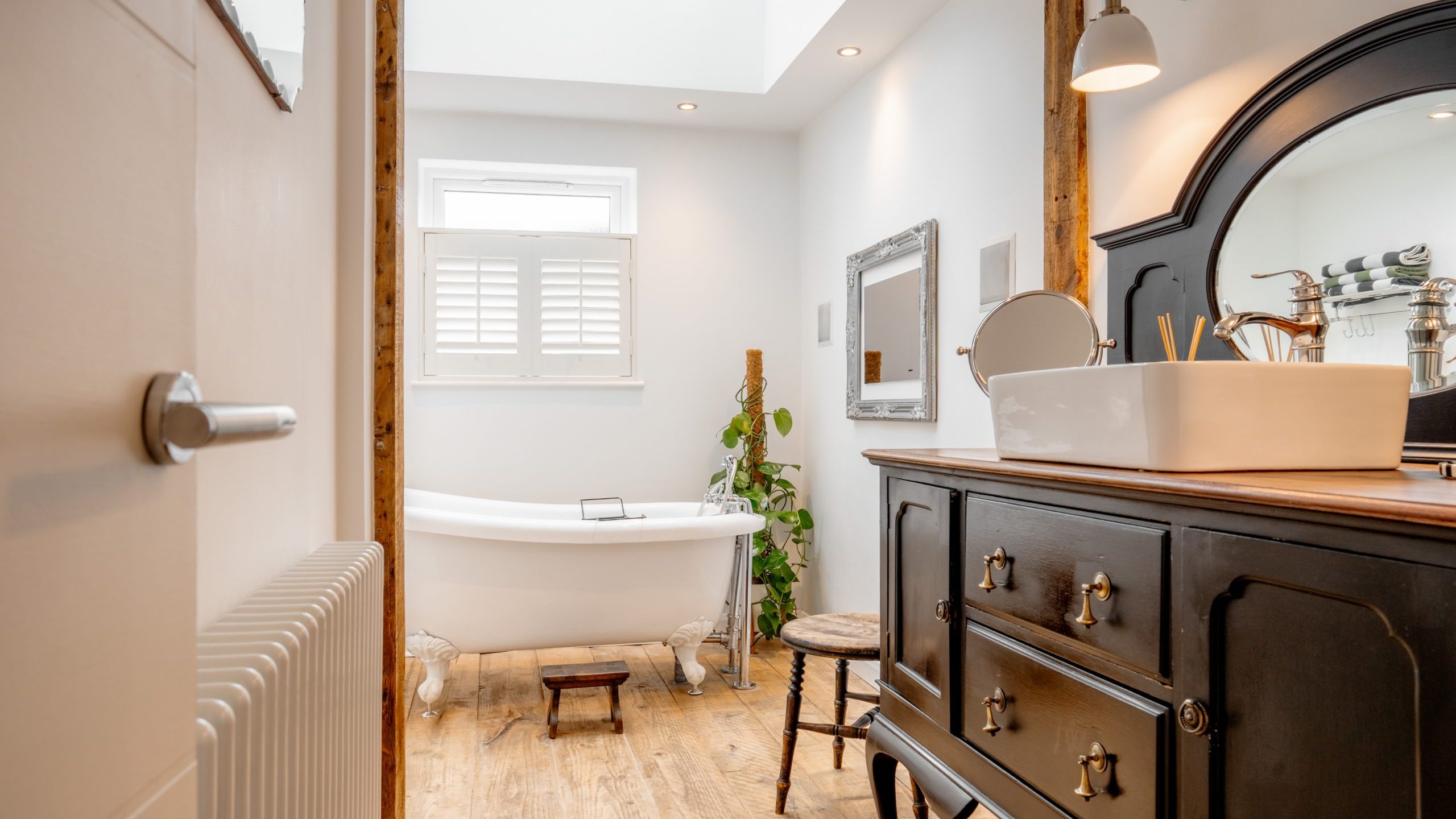 A cozy bathroom with a rustic wooden floor, featuring a freestanding clawfoot bathtub near a window. A black vanity with a vessel sink and round mirror is on the right. Soft lighting and a small plant create a warm ambiance.
