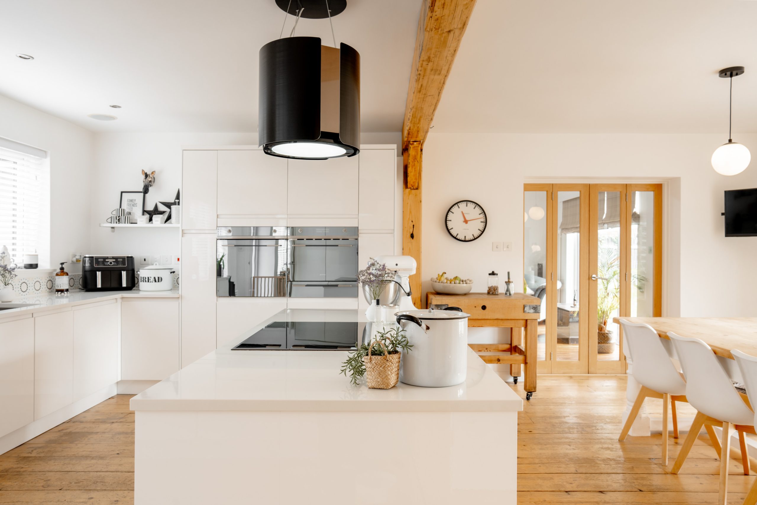 A modern kitchen with white cabinets and appliances. A large black pendant light hangs over a kitchen island. A rustic wooden beam and dining table with white chairs add warmth. A round clock and light wood flooring complete the space.