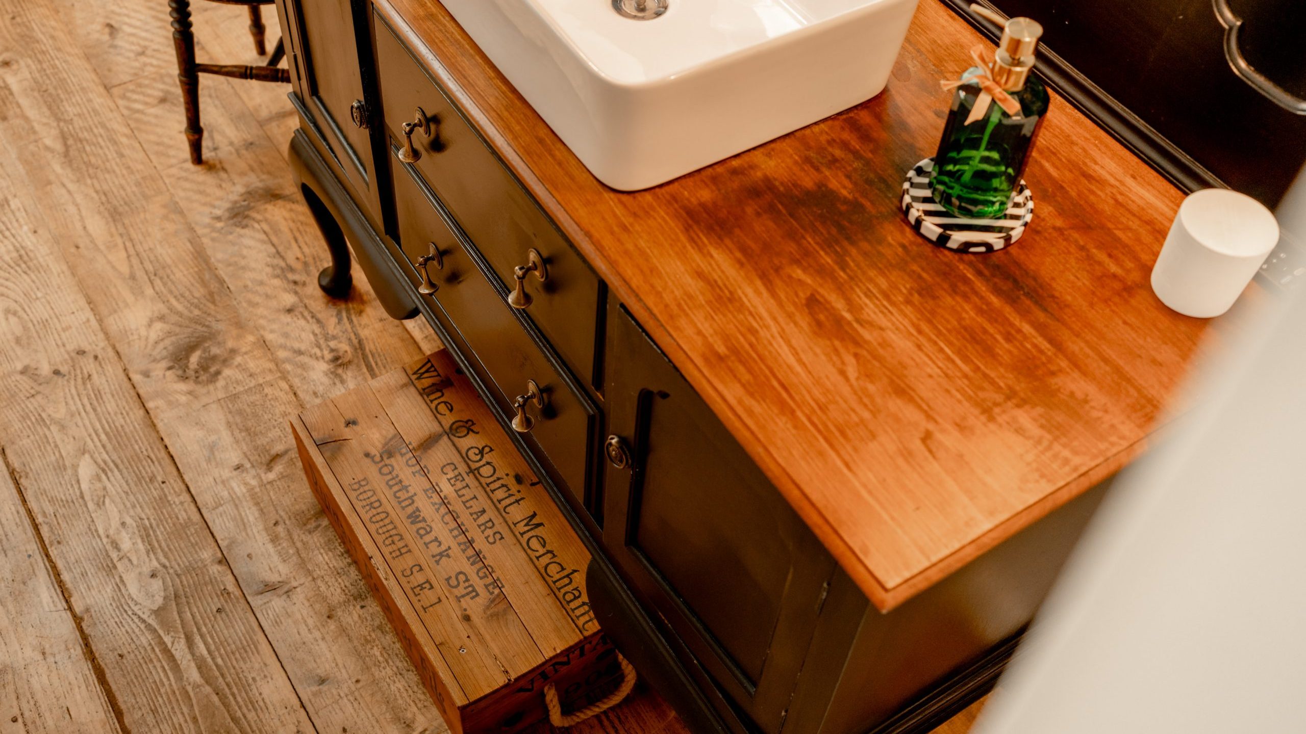 A wooden bathroom vanity with a white rectangular sink and a soap dispenser on top. An open drawer reveals an antique wooden crate inside. The floor is made of rustic wooden planks, and part of a chair is visible in the background.