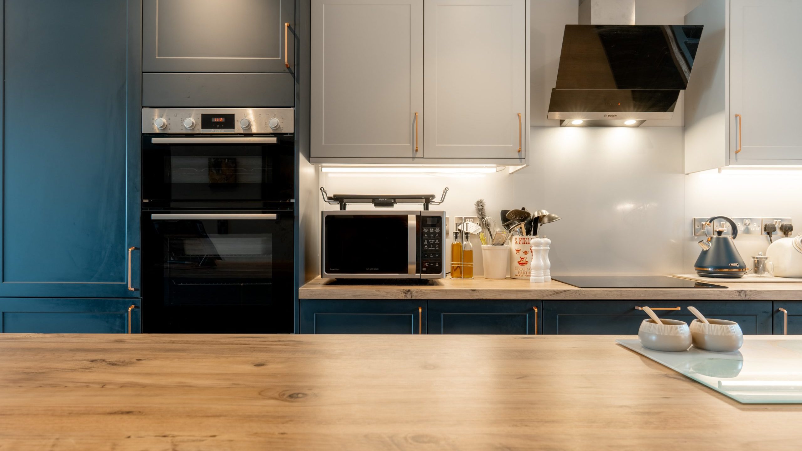 Modern kitchen with blue and white cabinets, a wooden countertop, built-in double oven, microwave, stovetop with black range hood, kettle, utensils, and jars neatly arranged on the counter.