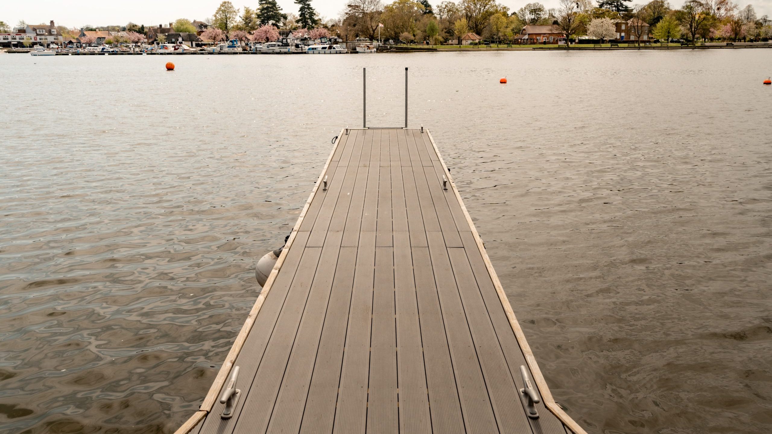 A wooden dock extends out over calm water toward the opposite shore, where houses and trees are visible. Orange buoys float in the water and the sky is partly cloudy.