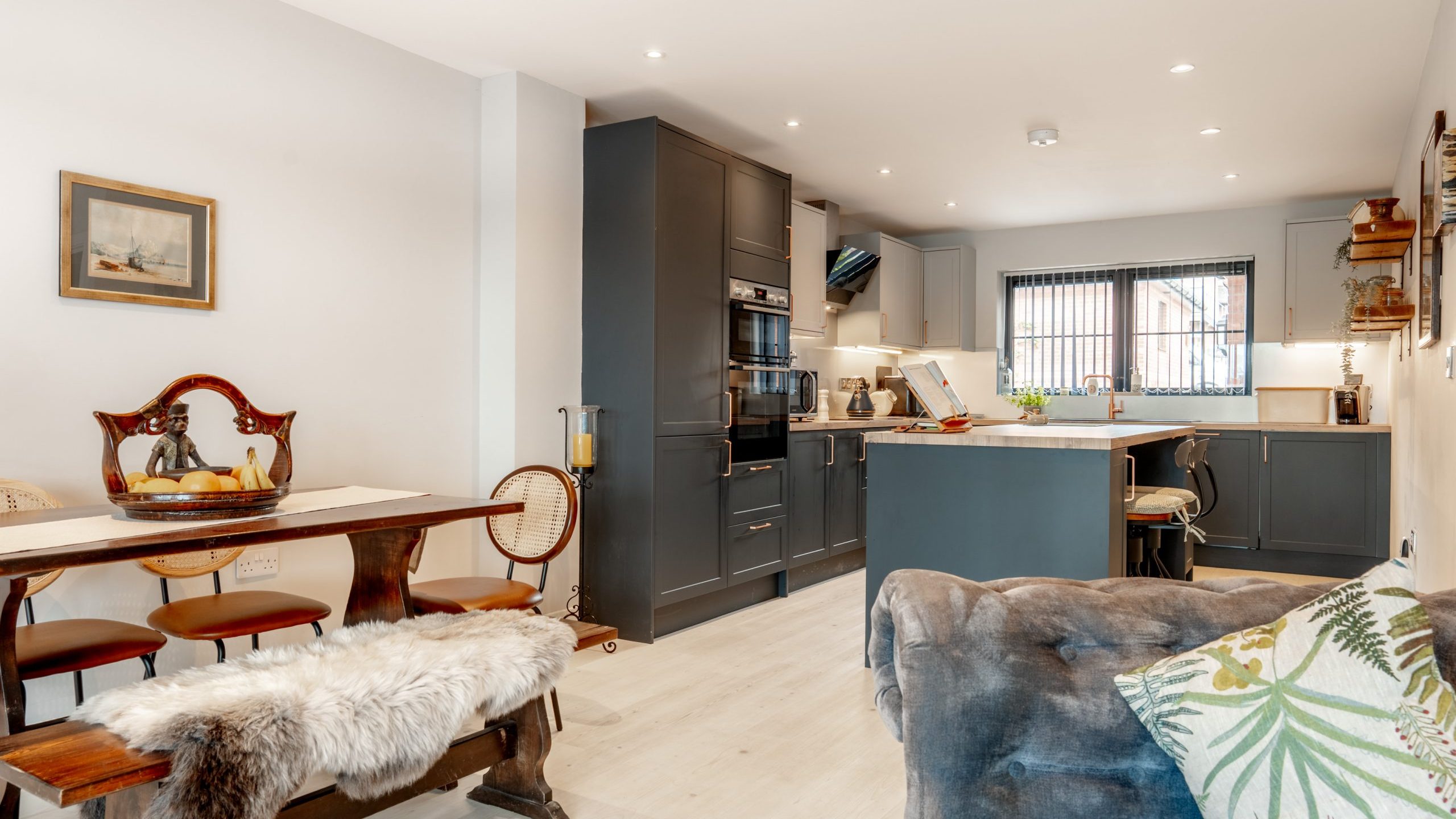Modern kitchen and dining area with gray cabinets, a wooden dining table with a bench and chairs, fur throw, cozy sofa with patterned pillows, and decor including a painting and fruit bowl; natural light streams in.