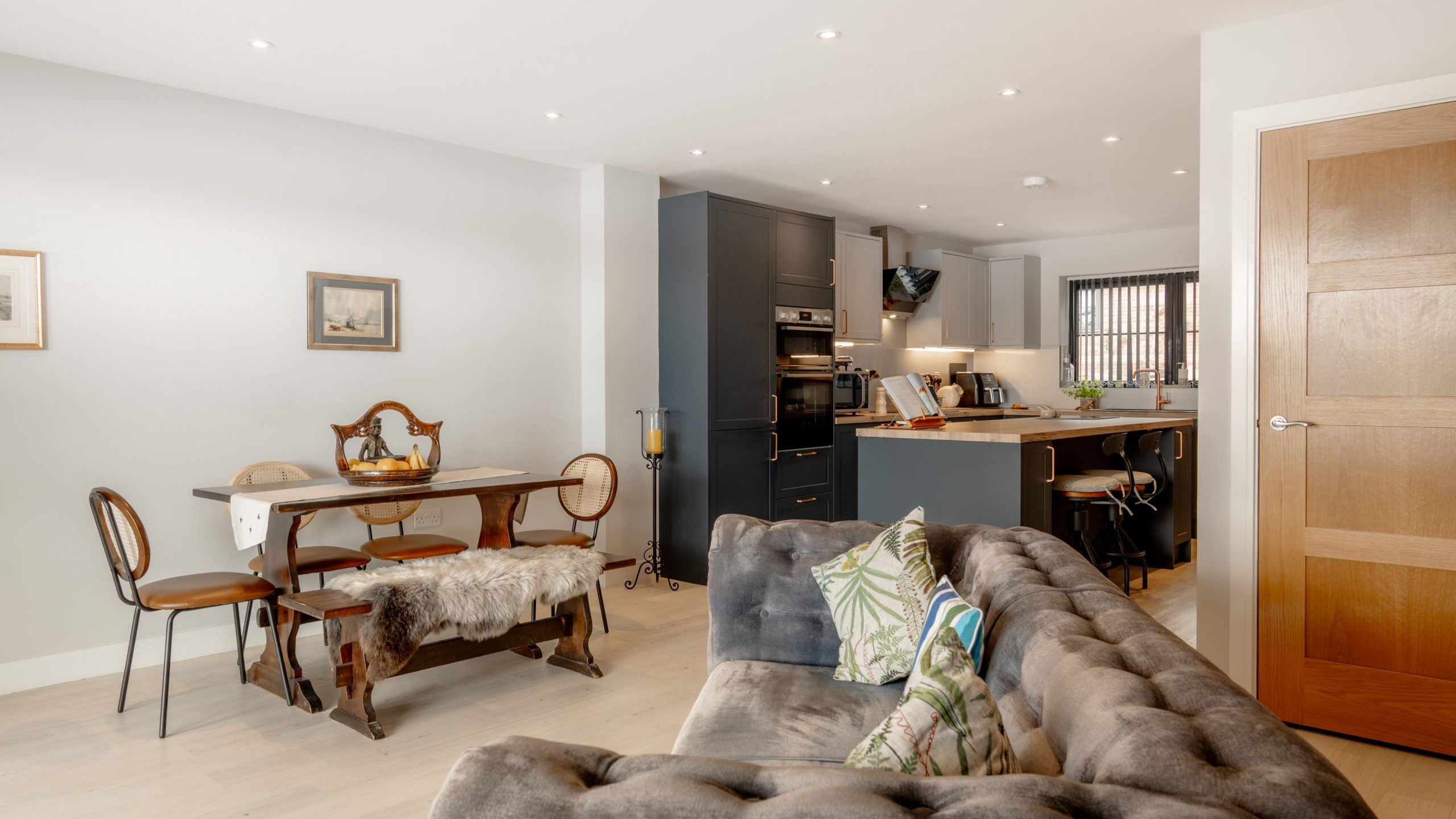 Open-plan living space with a gray tufted sofa, dining table with mixed chairs, and a modern kitchen featuring dark cabinets, built-in appliances, and a breakfast bar; neutral walls and wooden flooring.