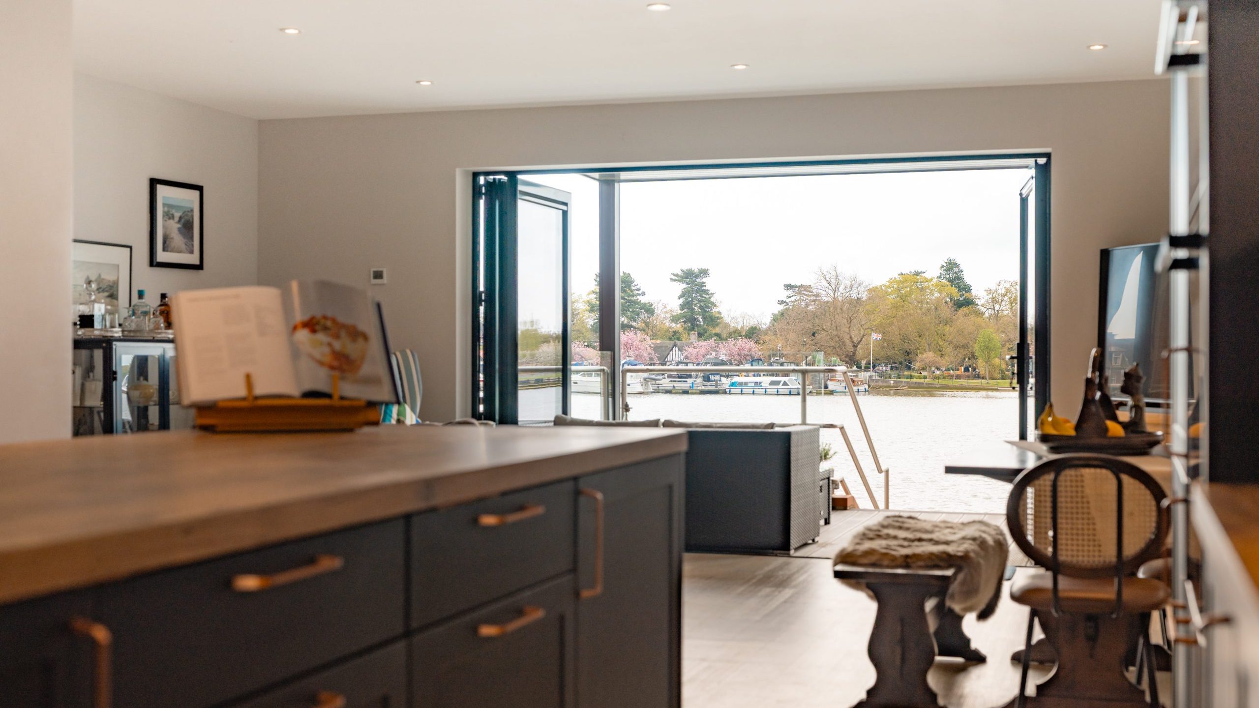 Modern kitchen and living area with open folding glass doors leading to a patio and a scenic riverside view with trees and boats outside. The space is bright, with wooden furniture and neutral tones.