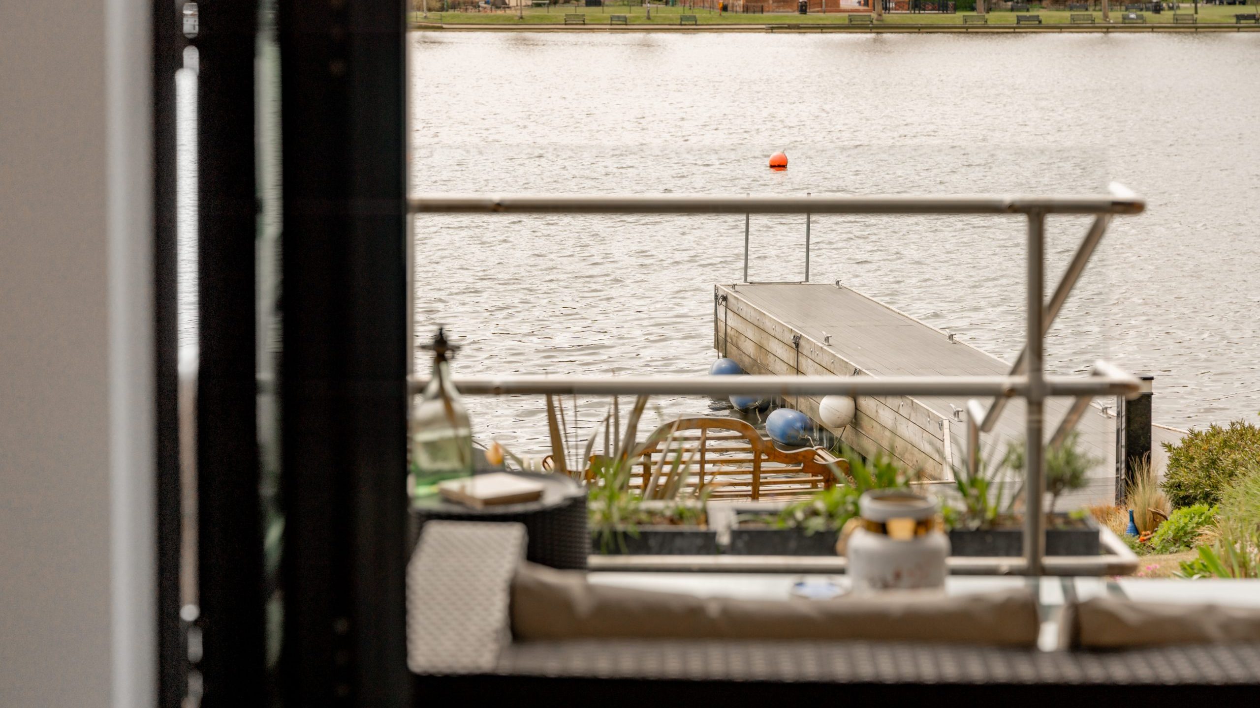 View from a balcony overlooking a dock extending into a calm river, with some outdoor furniture and potted plants in the foreground and houses with trees visible across the water.