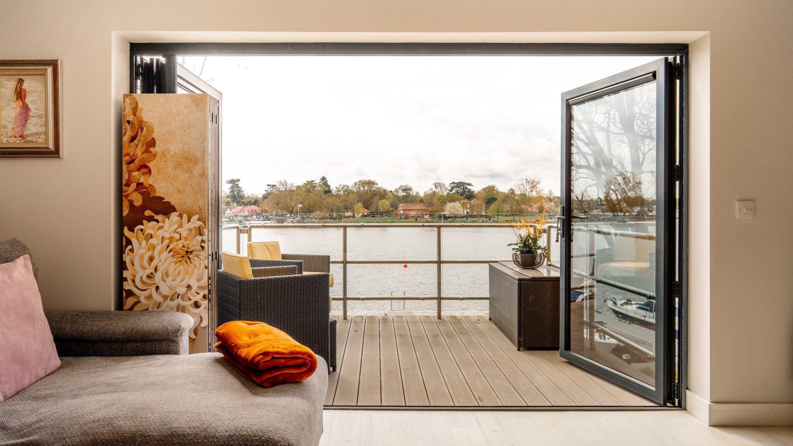 A cozy living room with a gray sofa and folded orange blanket opens to a wooden deck overlooking a calm river, with outdoor seating and trees visible across the water.
