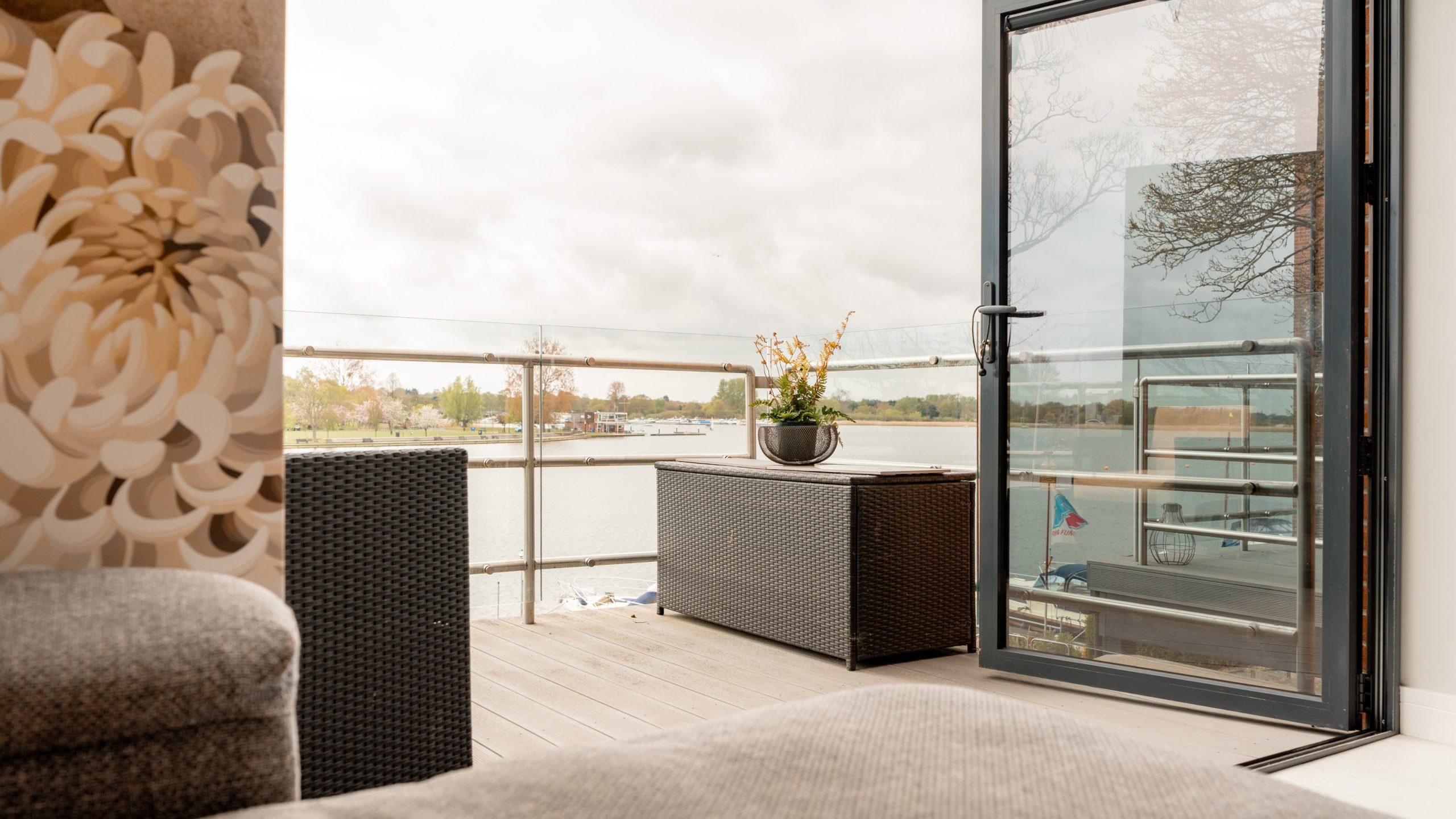 A cozy indoor space with a gray sofa opens onto a balcony with glass railing, wicker furniture, and potted flowers, overlooking a calm waterfront under a cloudy sky.