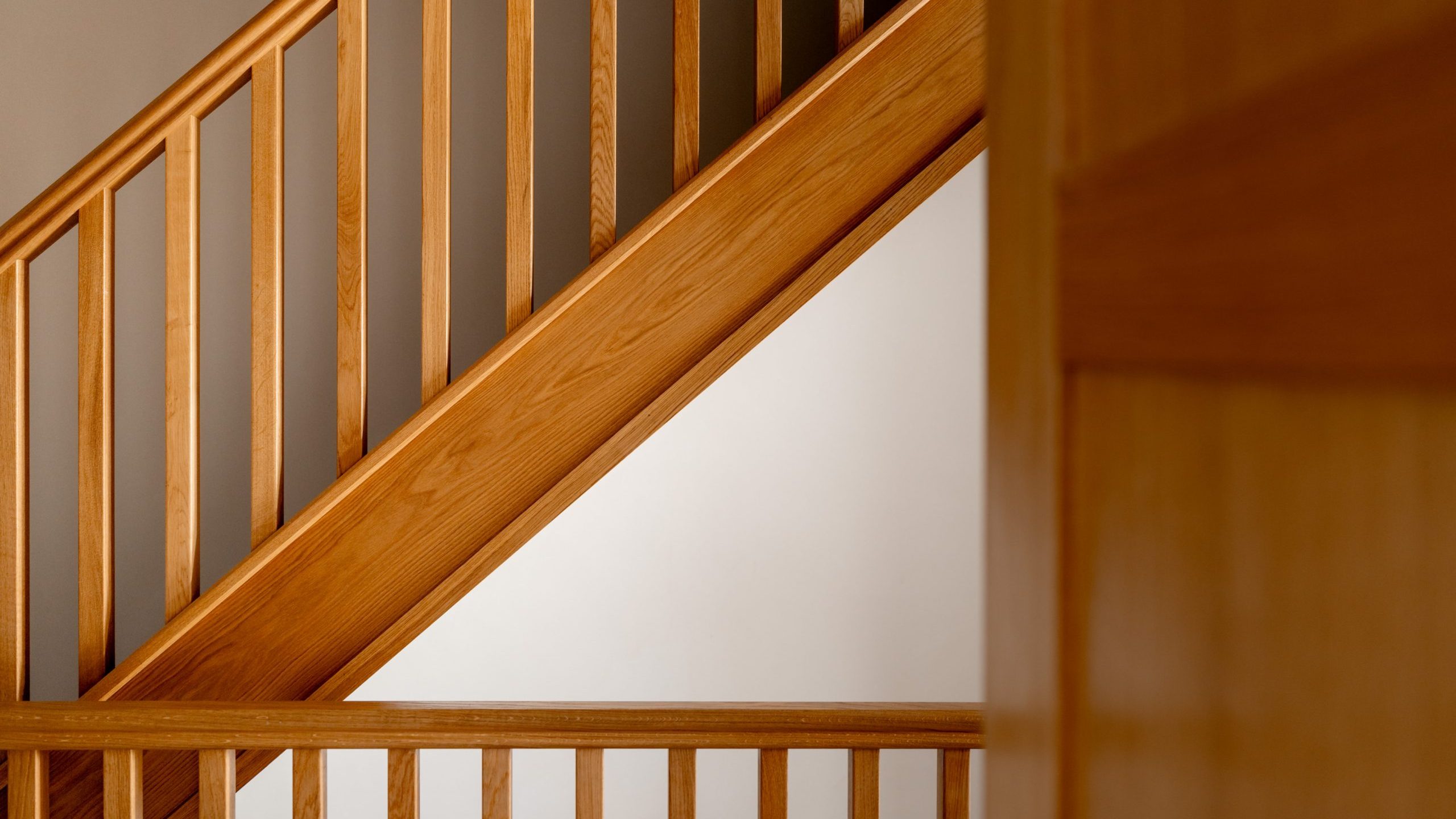 A close-up view of a wooden staircase with vertical railings and a handrail, showing warm brown wood tones and clean lines, with part of a wooden wall or panel in the foreground.