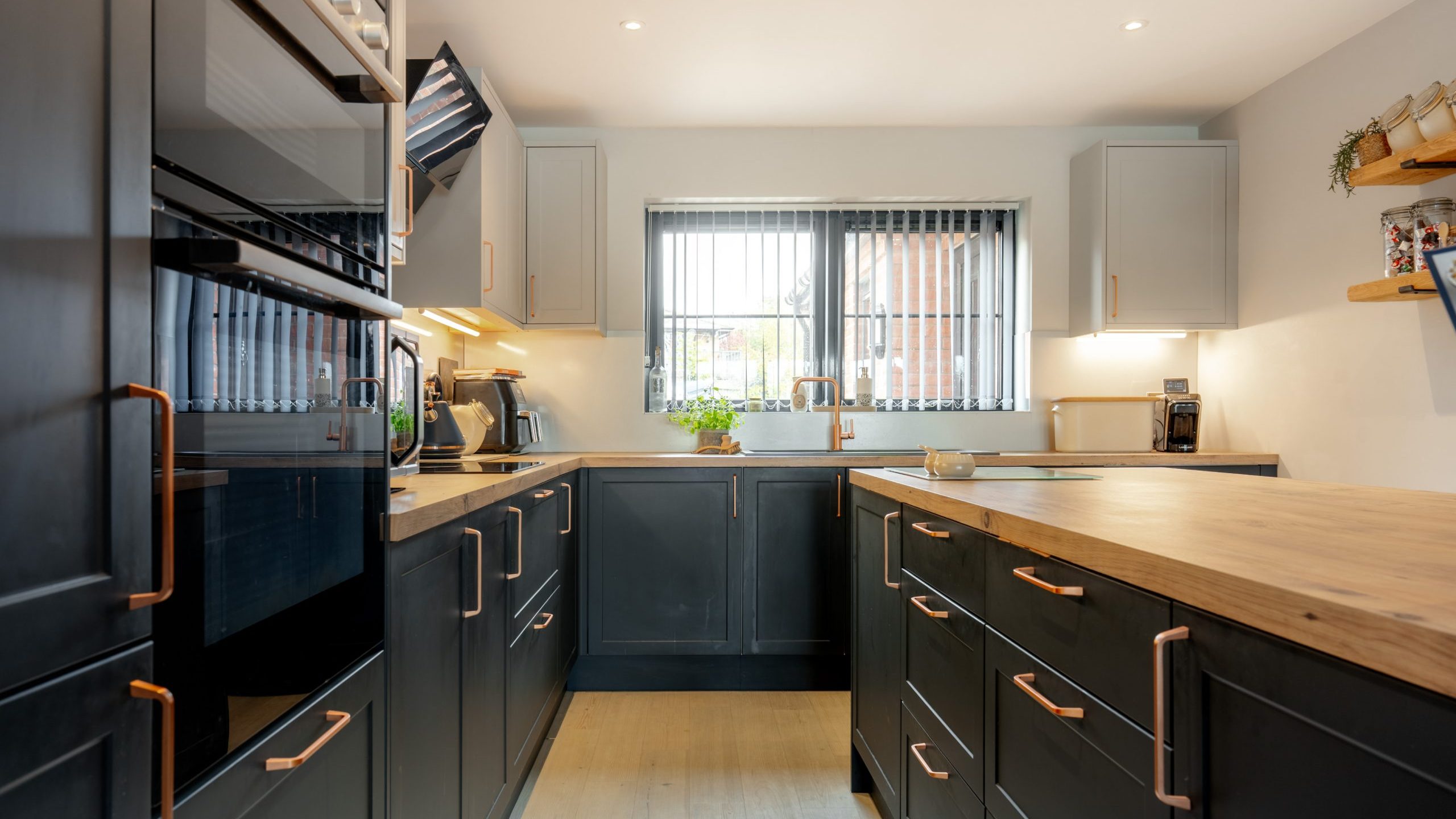 Modern kitchen with dark cabinets, wooden countertops, built-in oven, and open shelves. Large window with vertical blinds lets in natural light, and potted plant sits on the windowsill. The space is tidy and well-lit.