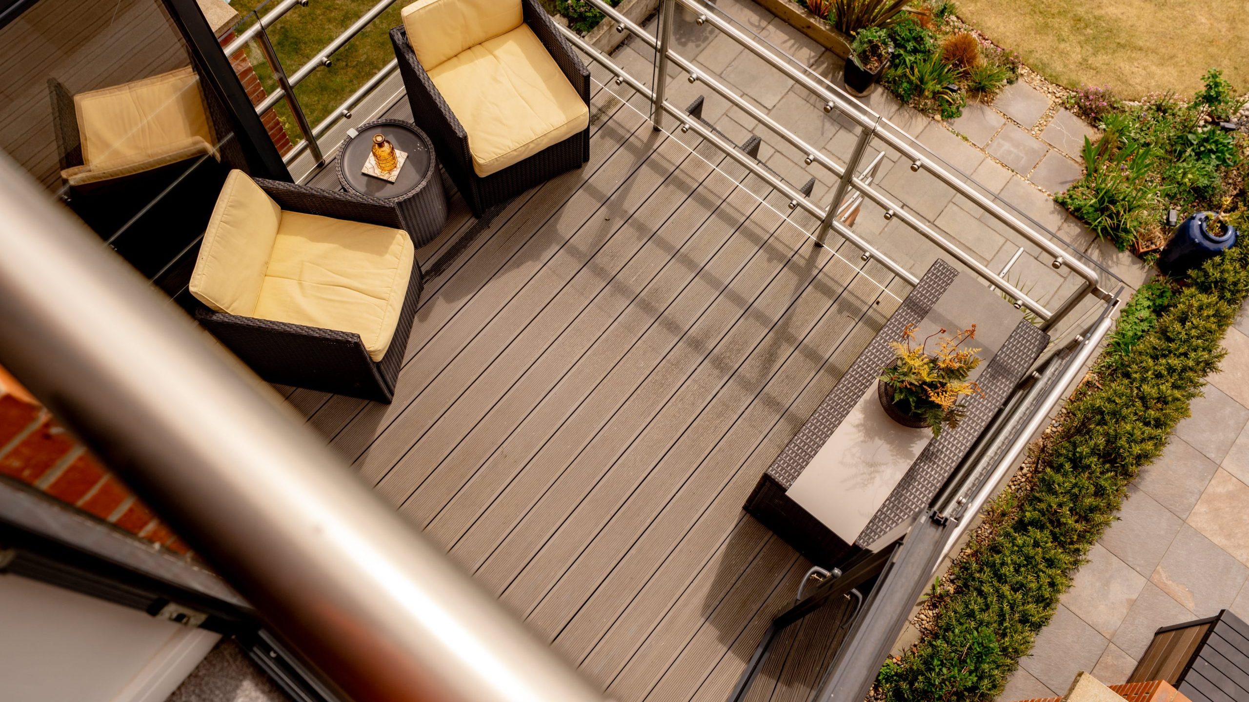 A top-down view of a modern patio with two cushioned chairs, a small table, potted plants, and a glass railing, overlooking a landscaped garden and stone walkway.