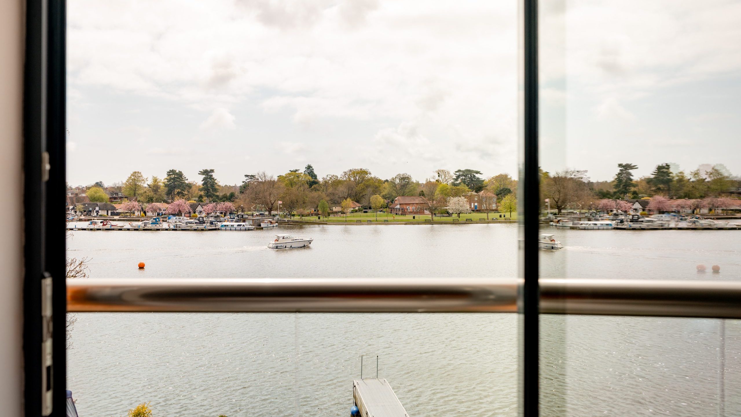 View through a glass window with a metal railing, overlooking a calm river with boats, a dock below, and houses with trees on the opposite riverbank under a cloudy sky.