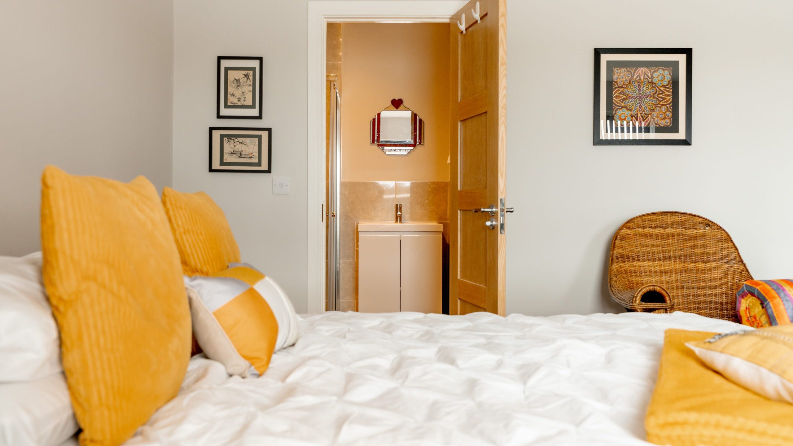 A cozy bedroom with a white bedspread and yellow pillows, facing an open wooden door that reveals a small bathroom with a sink and mirror. Art hangs on pale walls, and a wicker chair sits in the corner.