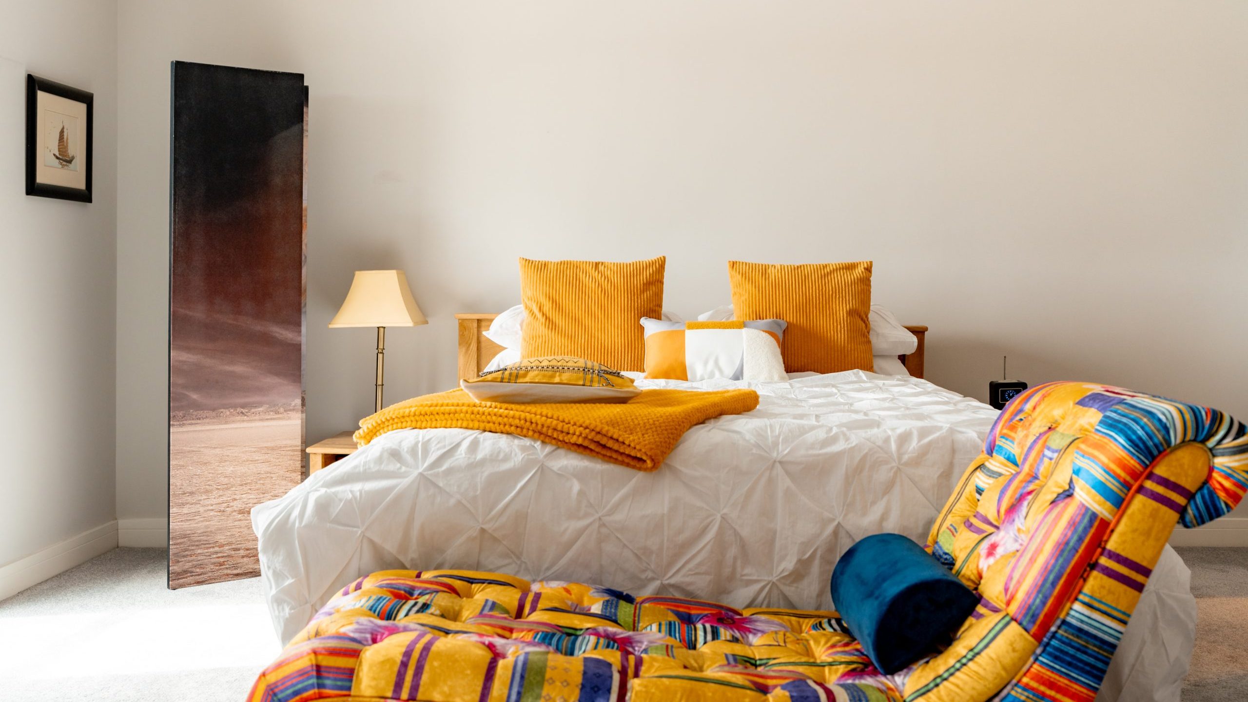 A cozy bedroom with a white bed, yellow pillows, a mustard throw blanket, a lamp, a decorative screen, and a colorful patterned chaise lounge with a blue pillow in the foreground.