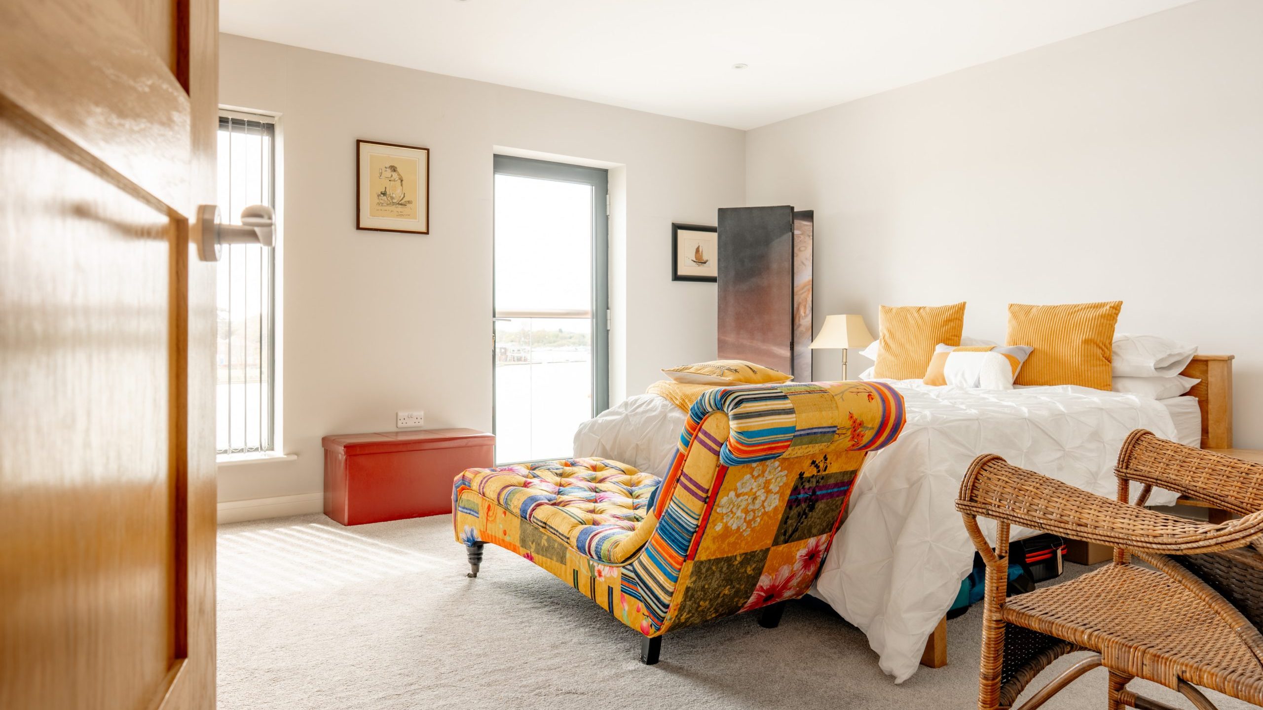 A bright bedroom with a double bed, white bedding, and yellow pillows. A colorful patchwork chaise lounge sits at the foot of the bed. There are wicker chairs, art on the walls, and a window letting in natural light.