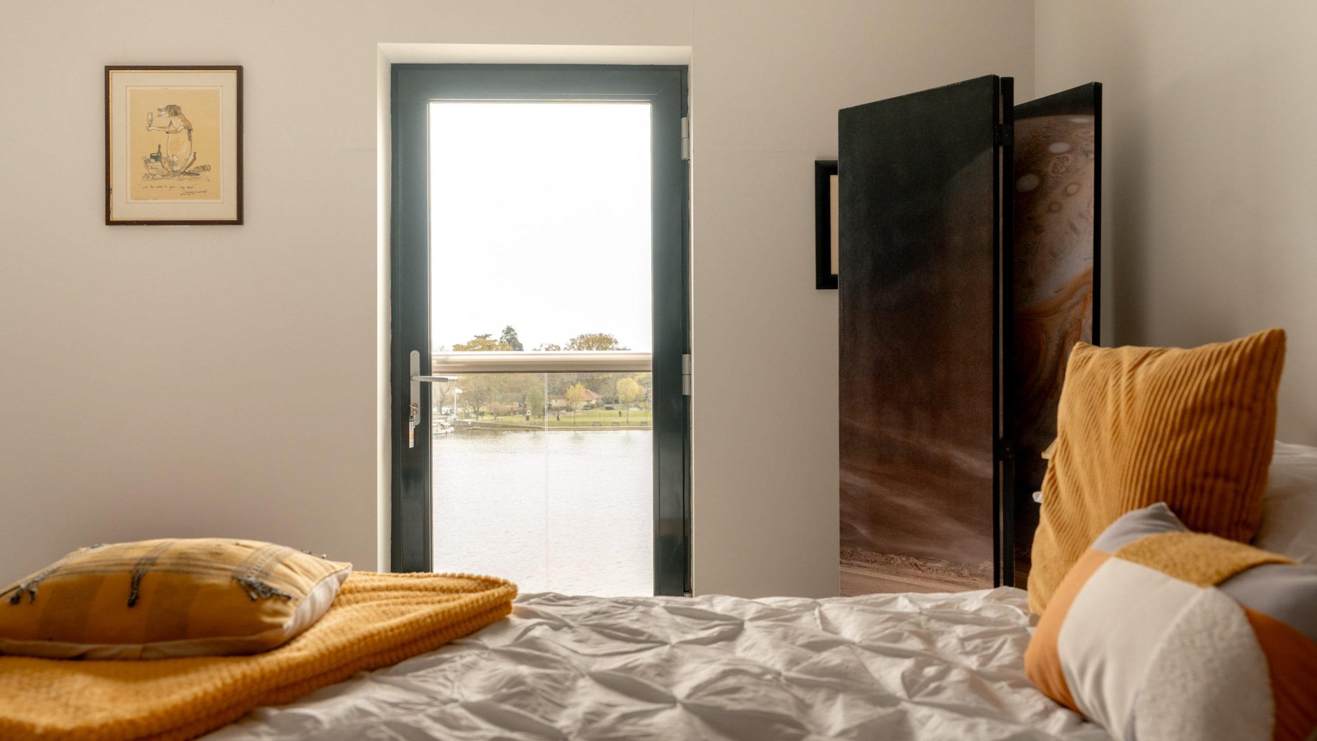 A cozy bedroom with a neatly made bed featuring yellow and white pillows and blanket, a framed picture on the wall, a dark-framed door with a window, and a decorative screen beside the door.