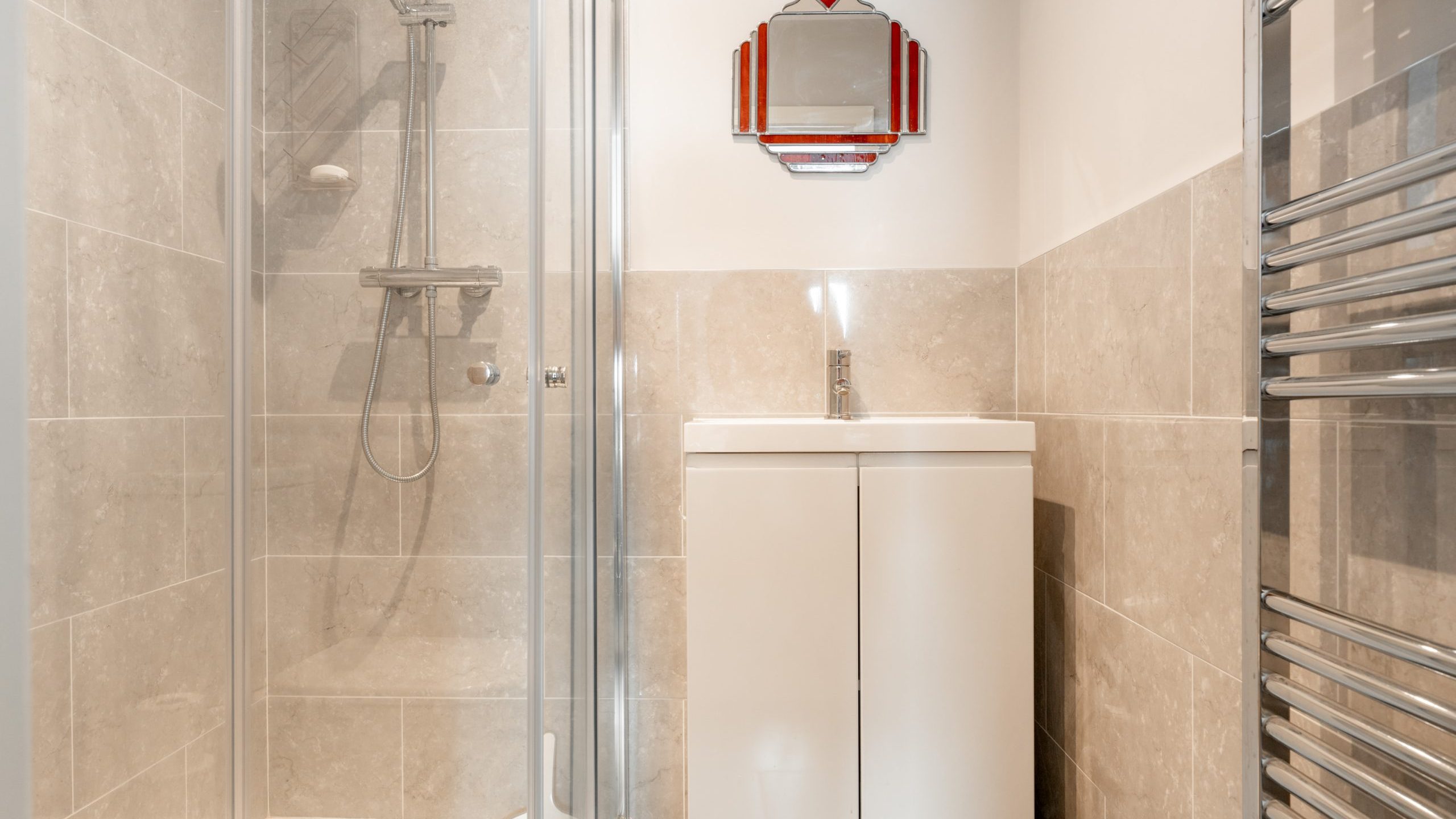 A modern bathroom with beige tiled walls, a glass shower enclosure on the left, a white vanity with a faucet in the center, a mirrored cabinet with a red heart detail above, and a chrome towel radiator on the right.