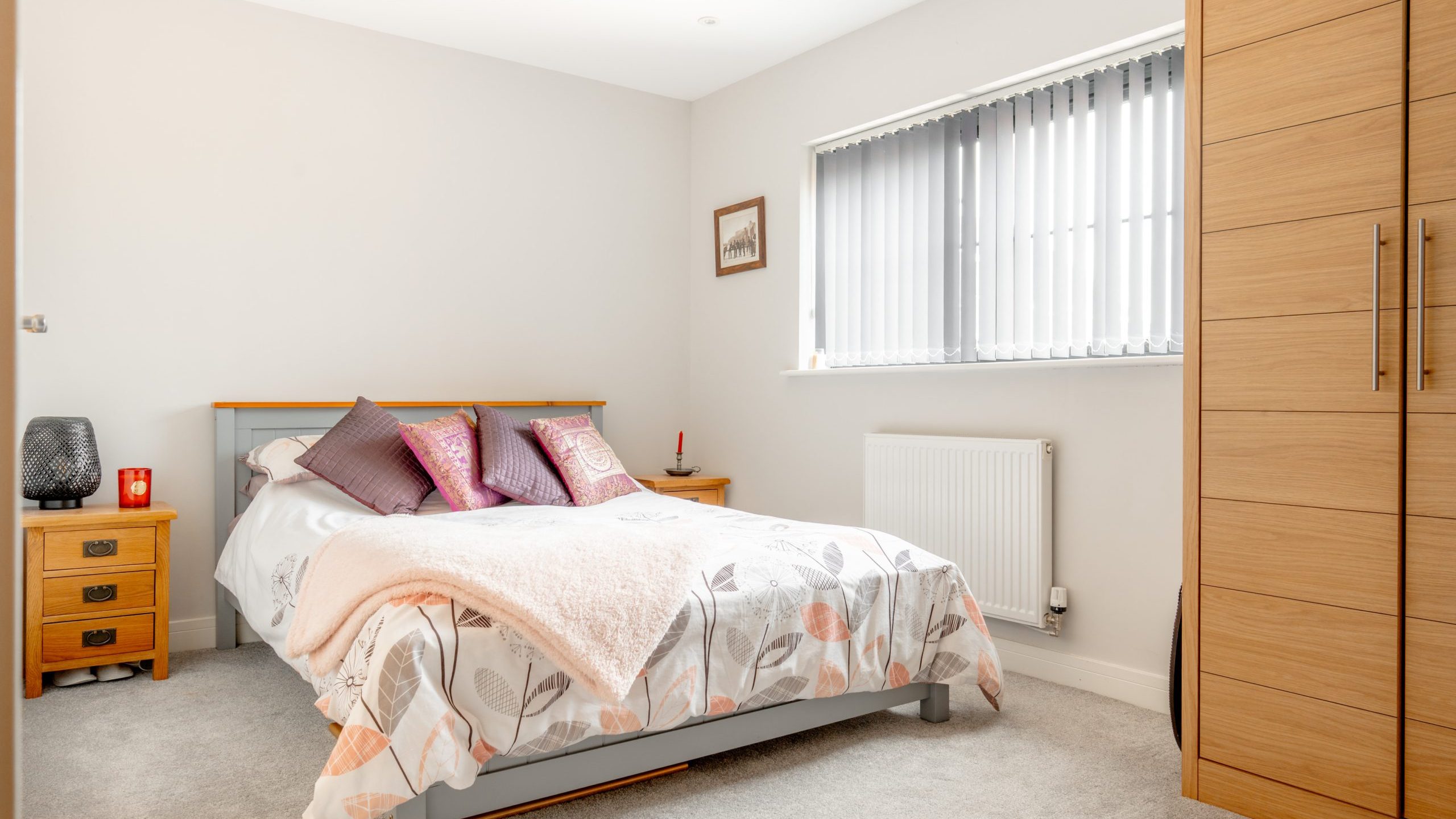 A bright, tidy bedroom with a double bed, floral bedding, purple and pink cushions, wooden nightstands, a tall wooden wardrobe, and a window with vertical blinds letting in natural light.