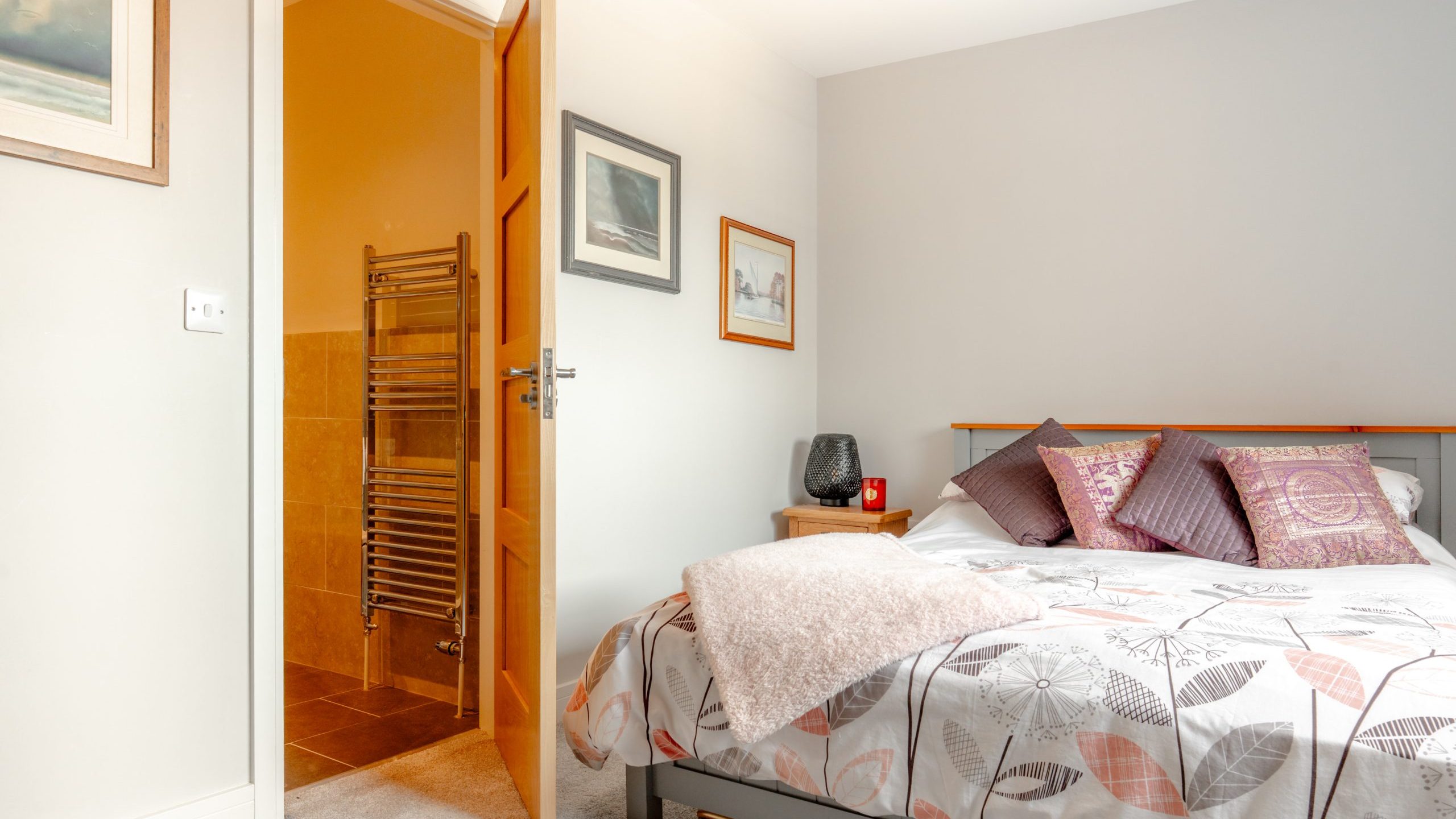A cozy bedroom with a double bed, patterned bedding, and decorative pillows. An open door reveals a bathroom with tan tiles and a heated towel rail. Framed art hangs on the light gray walls.
