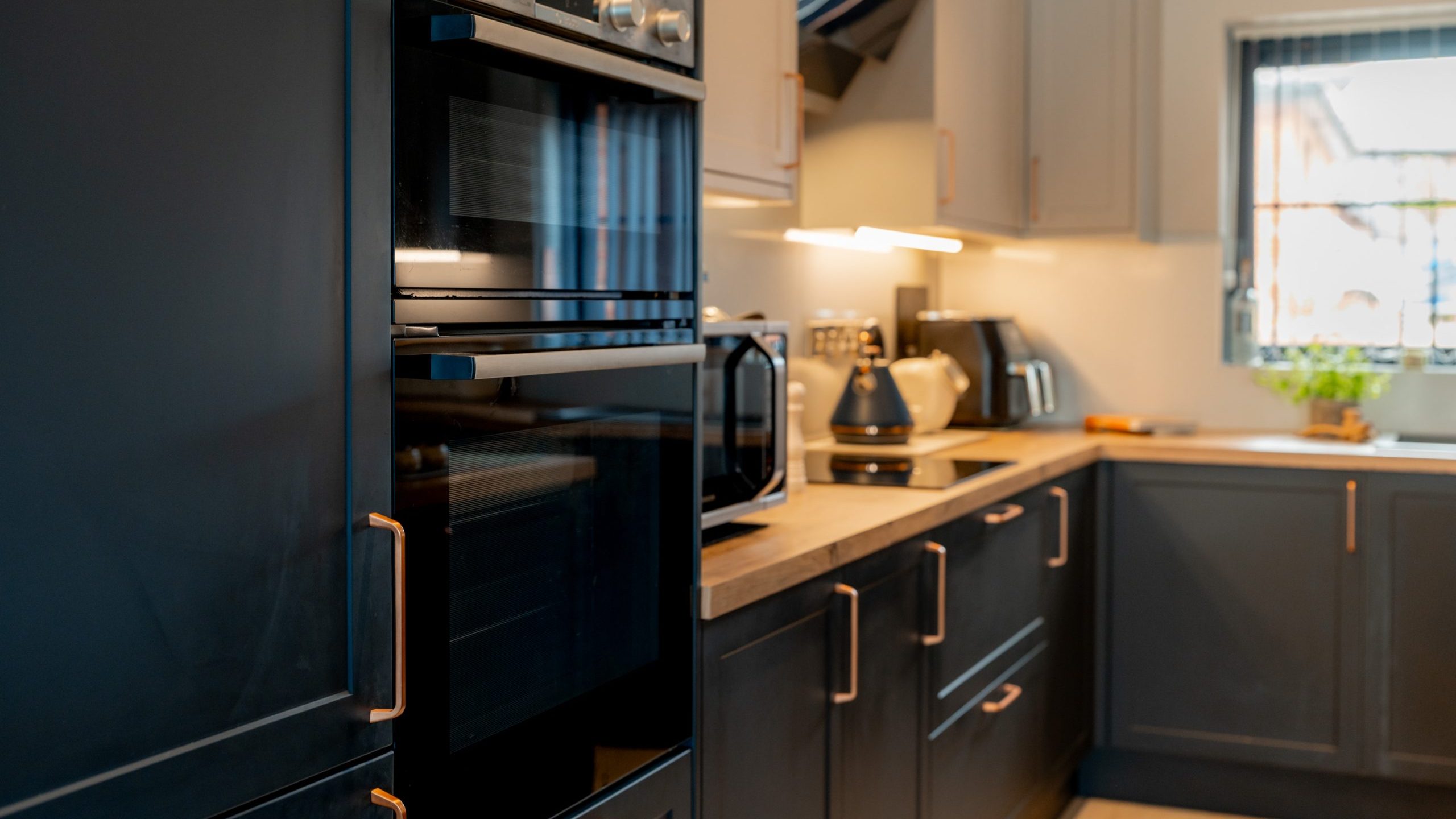 Modern kitchen with dark cabinets, built-in double oven, wooden countertops, electric stove, microwave, kettle, toaster, and a window letting in natural light.