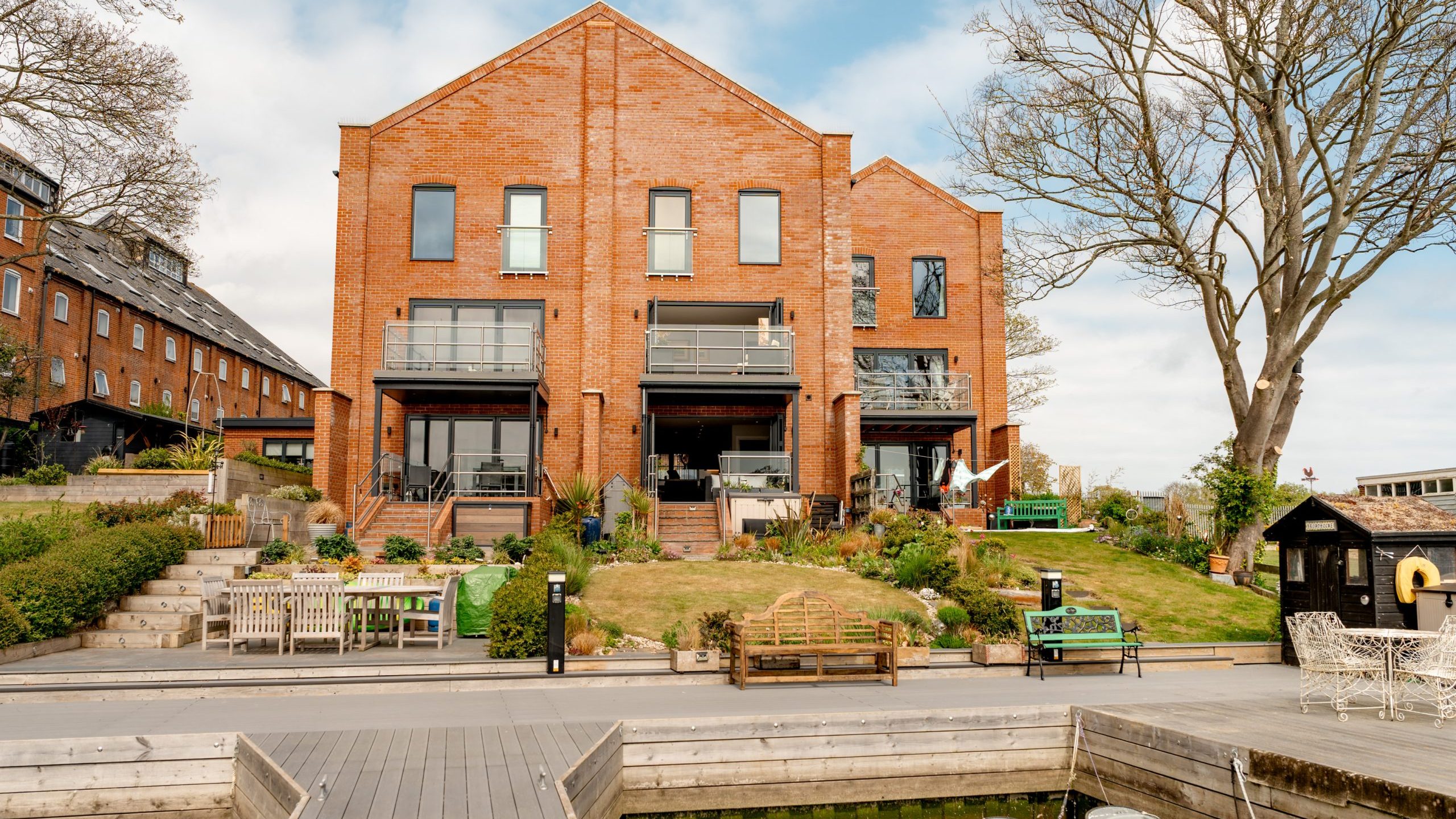 Two modern red-brick townhouses with large windows and balconies face a waterfront, surrounded by landscaped gardens, outdoor seating, and a wooden dock extending into the water.